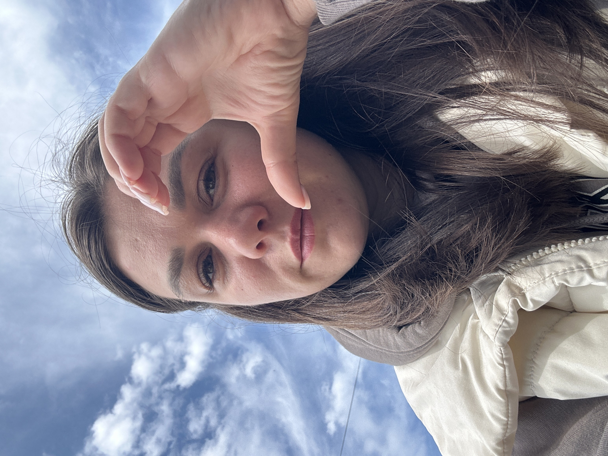 The image is a close-up, selfie-style shot of a young woman against a bright blue sky with wispy white clouds. She has long, dark brown hair and is wearing a light-colored puffer jacket over a hooded garment. Her right hand is positioned in front of her face, with her thumb and index finger forming a heart shape. The photo appears to have been taken outdoors, possibly in a park or open area, as suggested by the sky and the slight blur in the background, indicative of a shallow depth of field. There are no discernible landmarks that would indicate the specific location in Chaikovsky, Russia, or any other city. The focus is entirely on the woman and her gesture.
