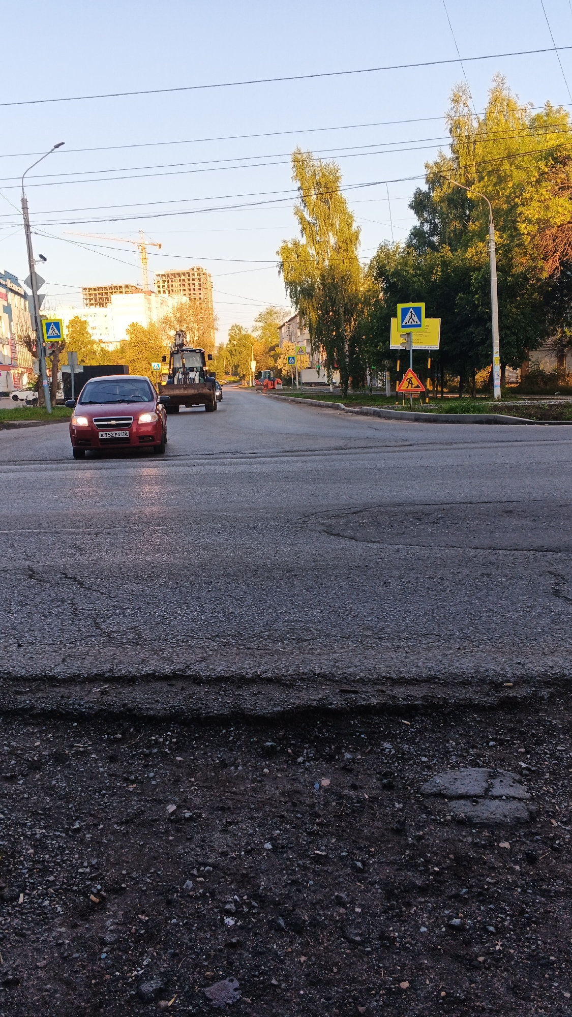 The image depicts a street scene in Izhevsk, Russia, likely in the morning or late afternoon due to the low angle of the sun casting long shadows and illuminating the trees from the side. The road is paved but shows signs of wear, with visible cracks and some sections appearing to be under repair or in disrepair, particularly in the foreground.
In the foreground, a red car is driving on the road. Its headlights are on. Behind the red car, a large construction vehicle, possibly a backhoe or loader, is also on the road, moving in the same direction. Further down the road, another orange construction vehicle is visible.
Several pedestrian crossing signs are present, including a blue square sign with a white pedestrian symbol. A yellow triangular warning sign with a pedestrian symbol and an exclamation mark is also visible on the right side of the road.
On the left side of the street, there are buildings and trees. In the background, a tall, multi-story building under construction, with a crane nearby, is visible. On the right side of the street, mature trees with green and yellow foliage line the road. Power lines are strung across the sky. The sky is clear and appears to be a pale blue. The overall atmosphere is that of an active urban environment with ongoing construction and traffic.