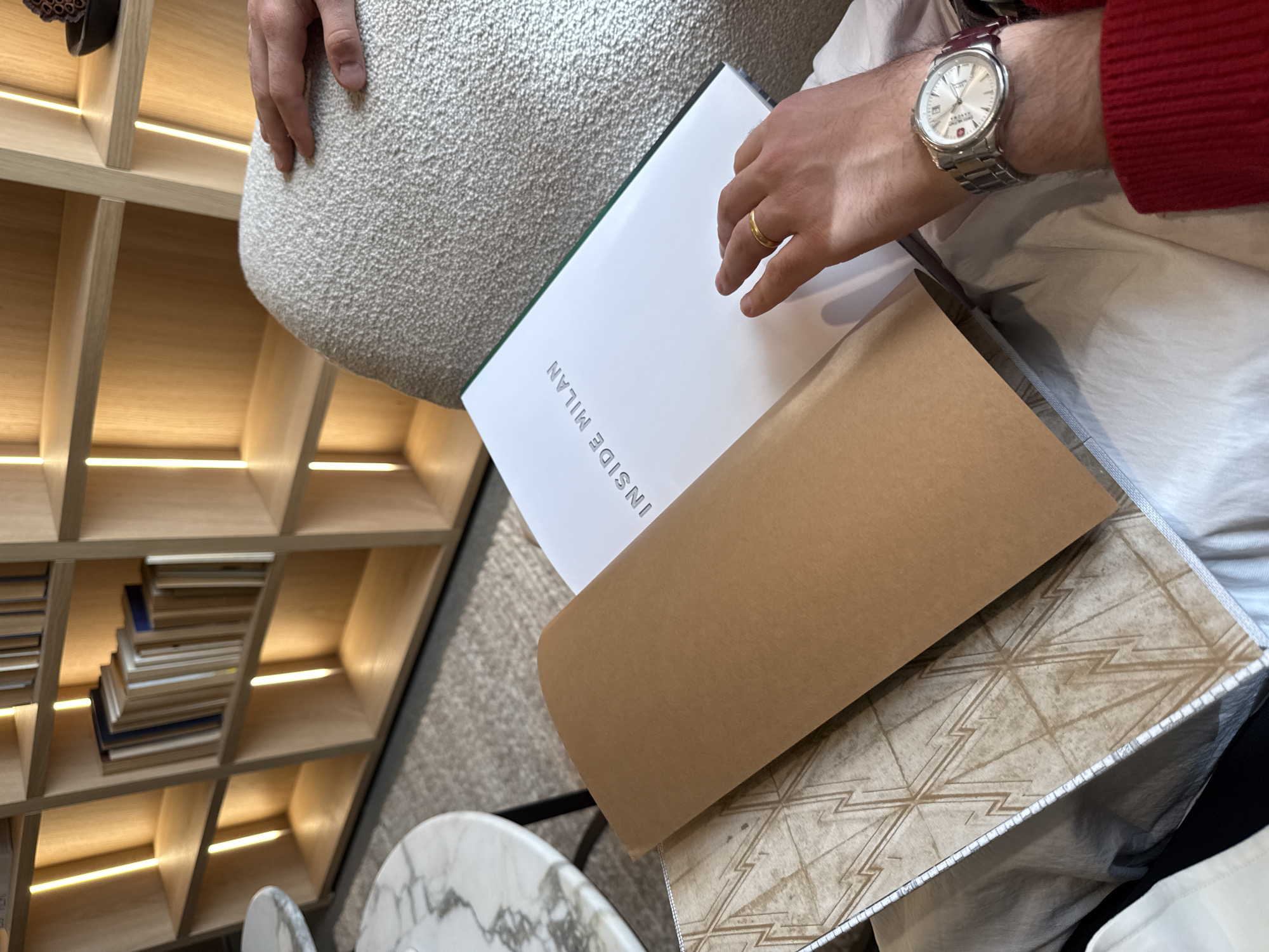 This media file captures an indoor scene in Milan, Italy, focusing on a person's hands and the materials they are reviewing. The person is seated, with their lap and lower torso covered by light-colored, textured fabric, likely a cushion or clothing. Their right hand, adorned with a simple gold band ring, rests on a white book with the title "INSIDE MILAN" prominently displayed in silver text. Their left wrist wears a silver-toned analog wristwatch featuring a metal link bracelet, a white dial with a date display, and a partially visible logo that includes a cross symbol.
Below the book, a large, plain brown piece of material, possibly cardstock or a folder, is visible, partially covering a patterned item with intricate, geometric zig-zag lines, suggesting it might be a fabric, tile, or wallpaper sample. In the background to the left, a modern wooden shelving unit with numerous square and rectangular compartments is visible. Each compartment is illuminated by a linear light strip, and some contain stacks of books or other decorative items. A fragment of a round table with a white and grey marble-like pattern is visible in the lower left corner. The floor appears to be covered with a light-colored, textured carpet. The scene suggests an activity of browsing or selecting design materials or references related to the city of Milan.