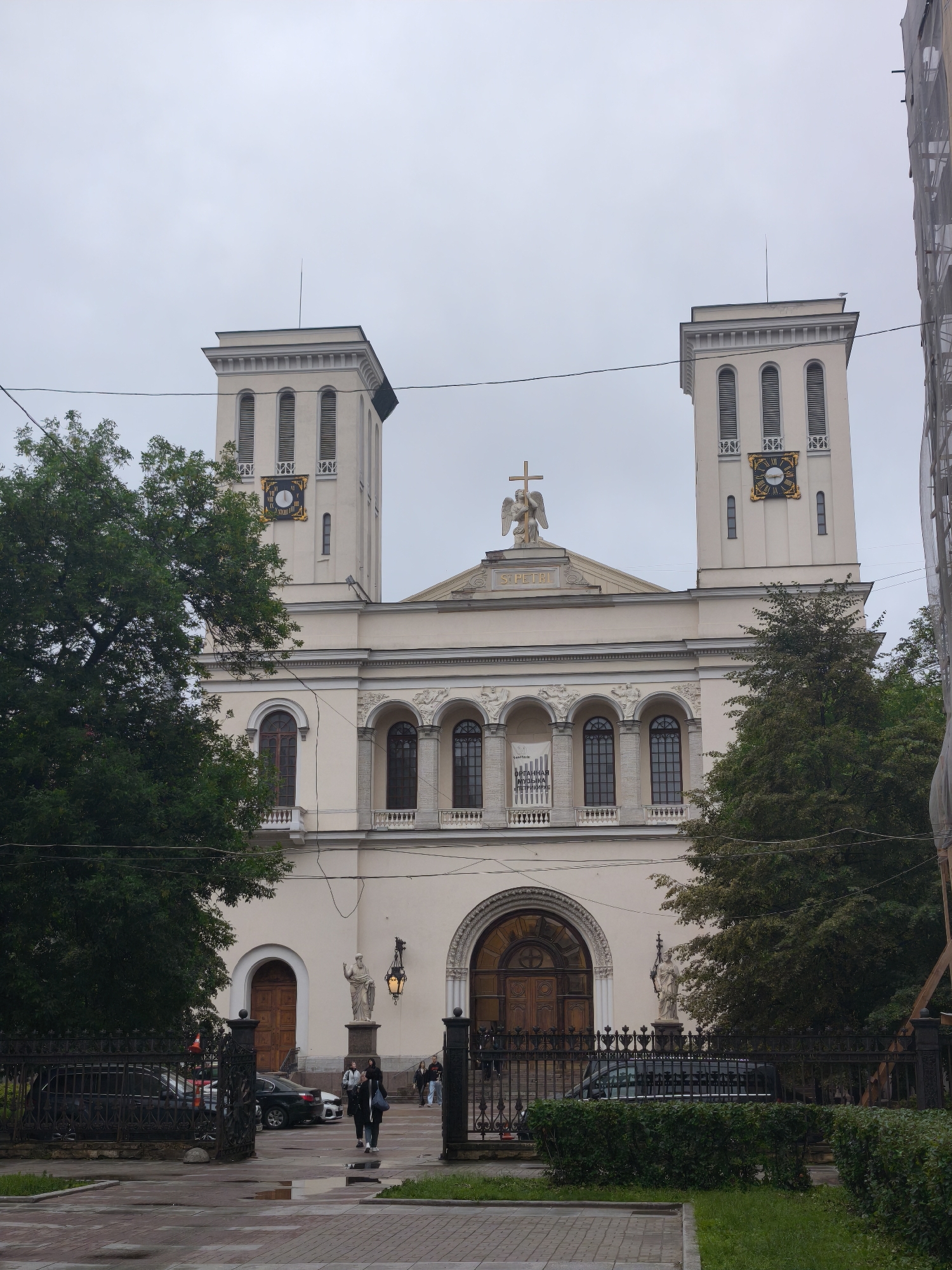 The image shows a wide, frontal view of the Saint Peter and Paul Lutheran Church in Saint Petersburg, Russia. The church is a light cream color with two prominent bell towers on either side of the main facade. Each tower features arched windows and decorative elements, and a clock face with Roman numerals is visible on the right tower. A statue of an angel holding a cross stands atop the central pediment of the church, with "S. PETRI" inscribed below it.
The central entrance of the church is a large, arched wooden door, flanked by arched windows and decorative sculptural elements. Two statues of saints stand on pedestals near the entrance, and an ornate lamppost hangs between them. A black metal fence encloses the church grounds, and several cars are parked behind it.
In the foreground, three people are walking on a paved walkway, suggesting a casual daytime scene. The sky is overcast, indicating cloudy weather. Lush green trees frame the sides of the church, partially obscuring the bell towers. A banner with Cyrillic text is visible on the facade of the church. The overall impression is one of a historic and serene religious building on a typical day in Saint Petersburg.