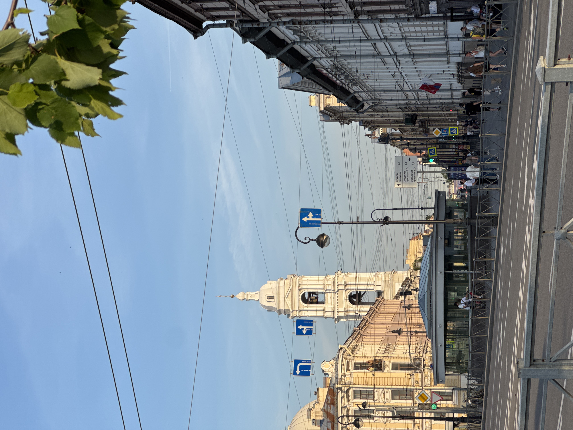 The image depicts a street scene in Saint Petersburg, Russia, likely in the historic center. A tall, white bell tower, a prominent architectural feature, dominates the mid-ground. Surrounding the tower are ornate buildings with classical detailing, characteristic of St. Petersburg's architecture. The street is wide and paved, with a pedestrian crossing in the foreground. Various traffic signs, including directional arrows and pedestrian crossing signs, are visible, indicating a busy urban environment. A modern glass-enclosed structure, possibly a metro station entrance or a public facility, is situated on the sidewalk. People are walking and cycling on the sidewalks and street. Overhead wires crisscross the sky, a common sight in older European cities. The sky is clear and blue, suggesting a pleasant day. A lush green tree branch hangs into the frame from the upper right corner, providing a touch of nature amidst the urban landscape. The overall impression is one of a vibrant city with a rich historical heritage.