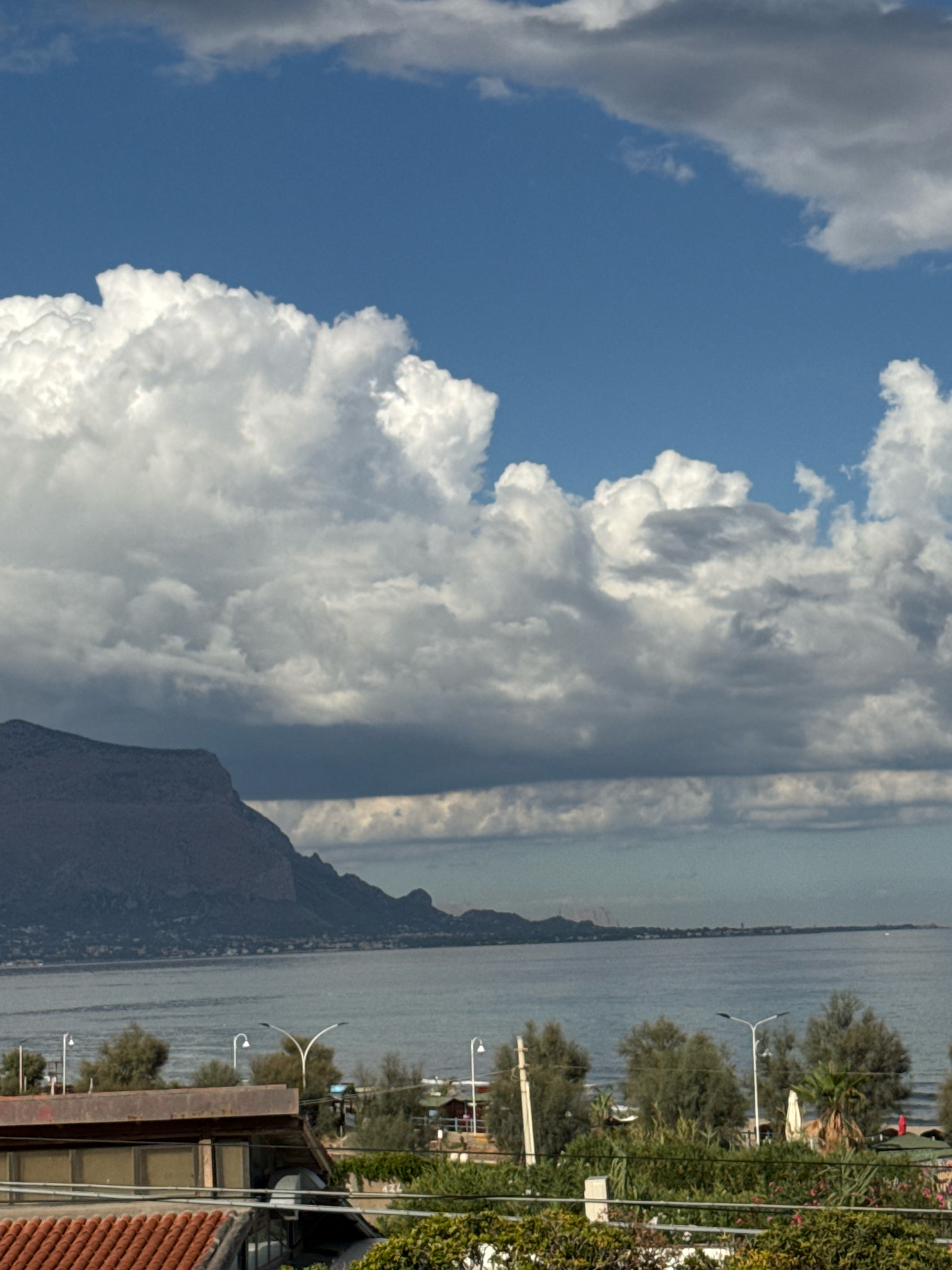 The image depicts a wide view of the coast of Capaci, Italy, under a dramatic sky filled with both bright white and dark gray clouds. The sea is calm and stretches to the horizon, where a distant coastline is visible with some low-lying buildings and what appear to be industrial structures. In the foreground, the roofs of buildings are visible, one with terracotta tiles and another with a brown, corrugated metal roof. Trees and vegetation dot the landscape between the buildings and the sea, with several streetlights also present. A large, imposing mountain range dominates the left side of the horizon, its rugged slopes rendered in shades of gray and brown. The overall impression is a coastal landscape, likely captured from an elevated vantage point, with a mix of natural beauty and human development.
