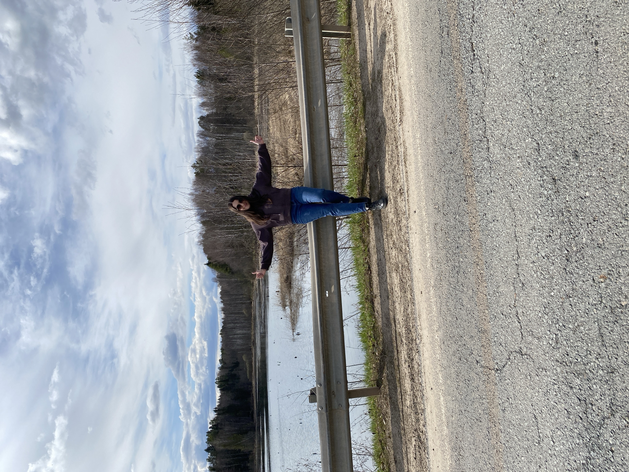 A young woman stands leaning against a guardrail on the side of a road. She has long brown hair and wears sunglasses, a dark jacket, and blue jeans. She has her arms outstretched. To her left, beyond the guardrail, is a body of water. On the opposite side of the water is a forest. The sky is overcast. The road appears to be paved with gray asphalt. The location is potentially Kamennyi Kliuch, Russia, judging by the question prompt.