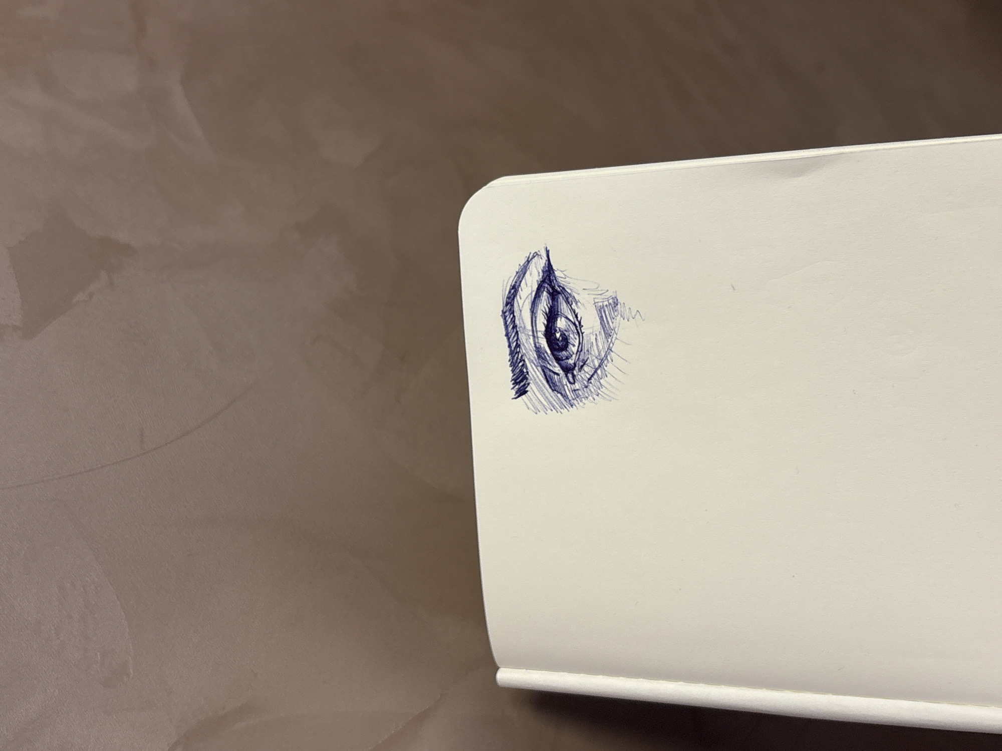 A close-up shot depicts a white, possibly plastic, surface adjacent to a wall with a textured, beige or light brown finish. On the white surface, a pen drawing of an eye is visible, executed in blue ink. The drawing shows details of the eye structure including eyelashes. The location is not discernible from the given image.
