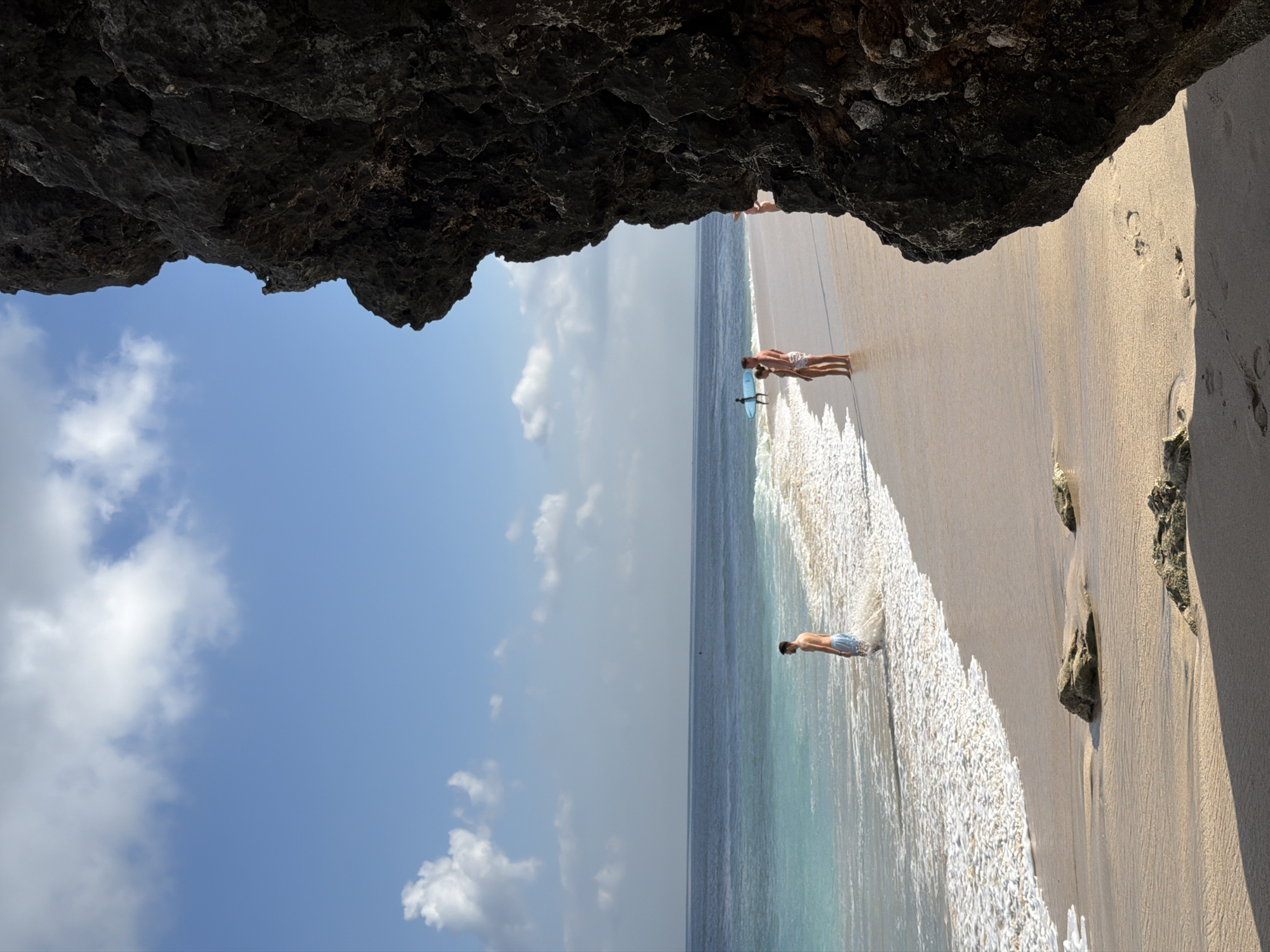 Captured from inside what appears to be a cave, the image shows a beach scene in Kuta Selatan, Indonesia. The sky, partially covered by clouds, contrasts with the dark, rocky ceiling of the cave structure framing the top of the shot. Below, the shoreline is visible, featuring light-colored sand and waves meeting the beach. Several individuals are on the beach: a man is standing in the water's edge, while further down the beach, two people stand beside a light-blue surfboard. Footprints dot the sandy beach.