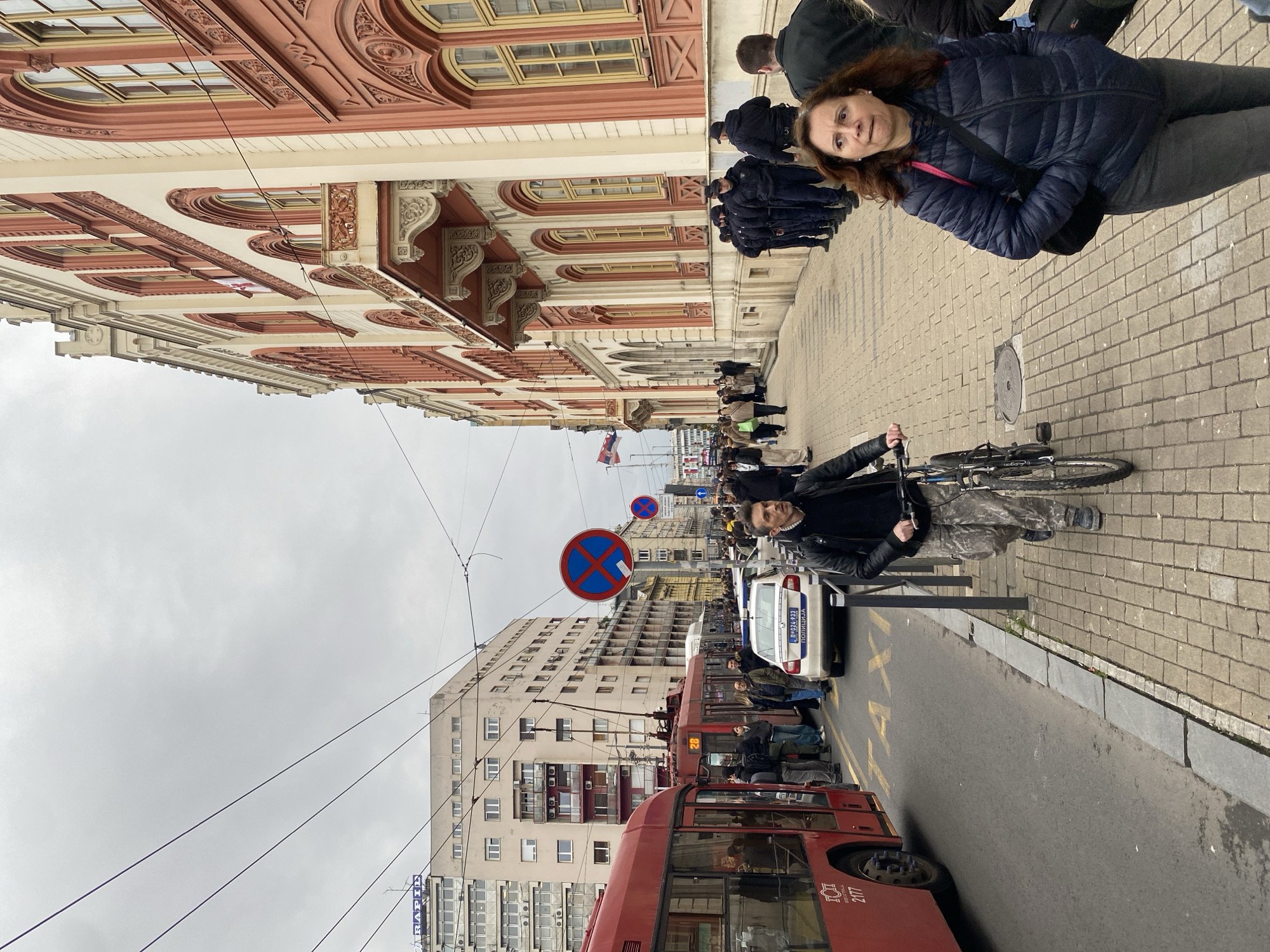 The image shows a street scene in Belgrade, Serbia. A red articulated bus is stopped on the street next to a white police car. On the sidewalk, a man on a bicycle is walking, and a woman in a blue jacket is walking in the opposite direction. A crowd of people is further down the sidewalk. A no-stopping sign is visible. Buildings line both sides of the street, with one side featuring ornate architectural details and the other a more utilitarian style. The sky is overcast. A Serbian flag is visible in the distance.