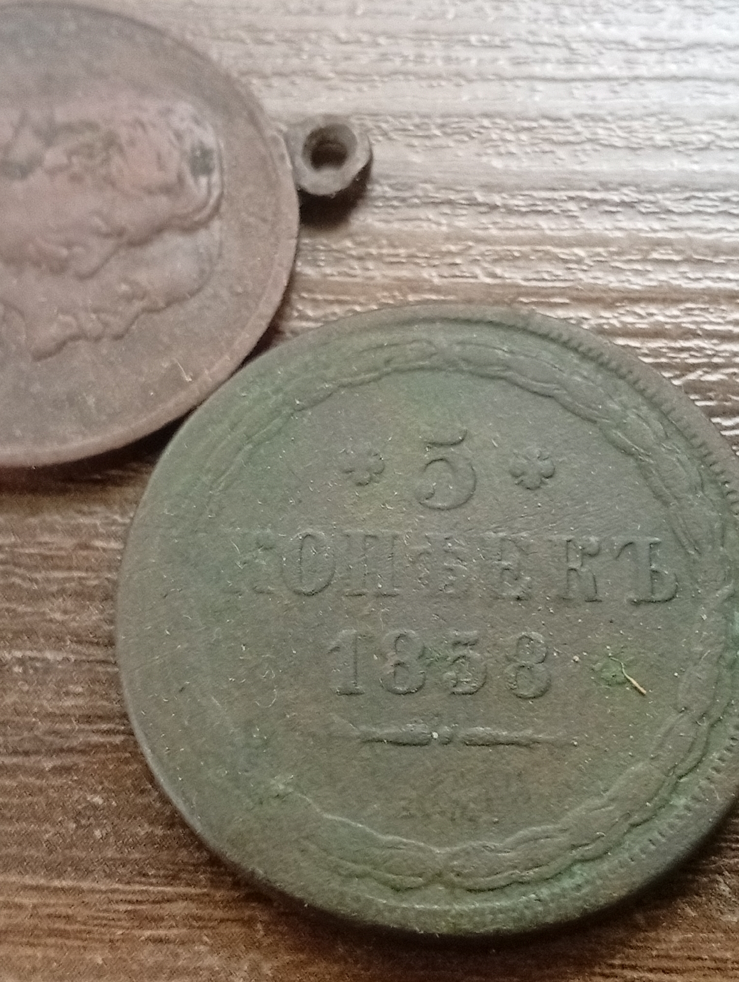 The image shows two old coins on a wooden surface. The coin in the foreground is round and appears to be made of copper, with the inscription "5 КОПЄЕКЪ 1858" visible. The coin in the background is also round and appears to be made of a lighter metal, with a profile of a person on it. The wooden surface has a wood grain pattern and is light brown in color. The scene is lit from above, casting soft shadows. No people are visible in the image. The location of Varputėnai, Lithuania, is not discernible from the image itself.