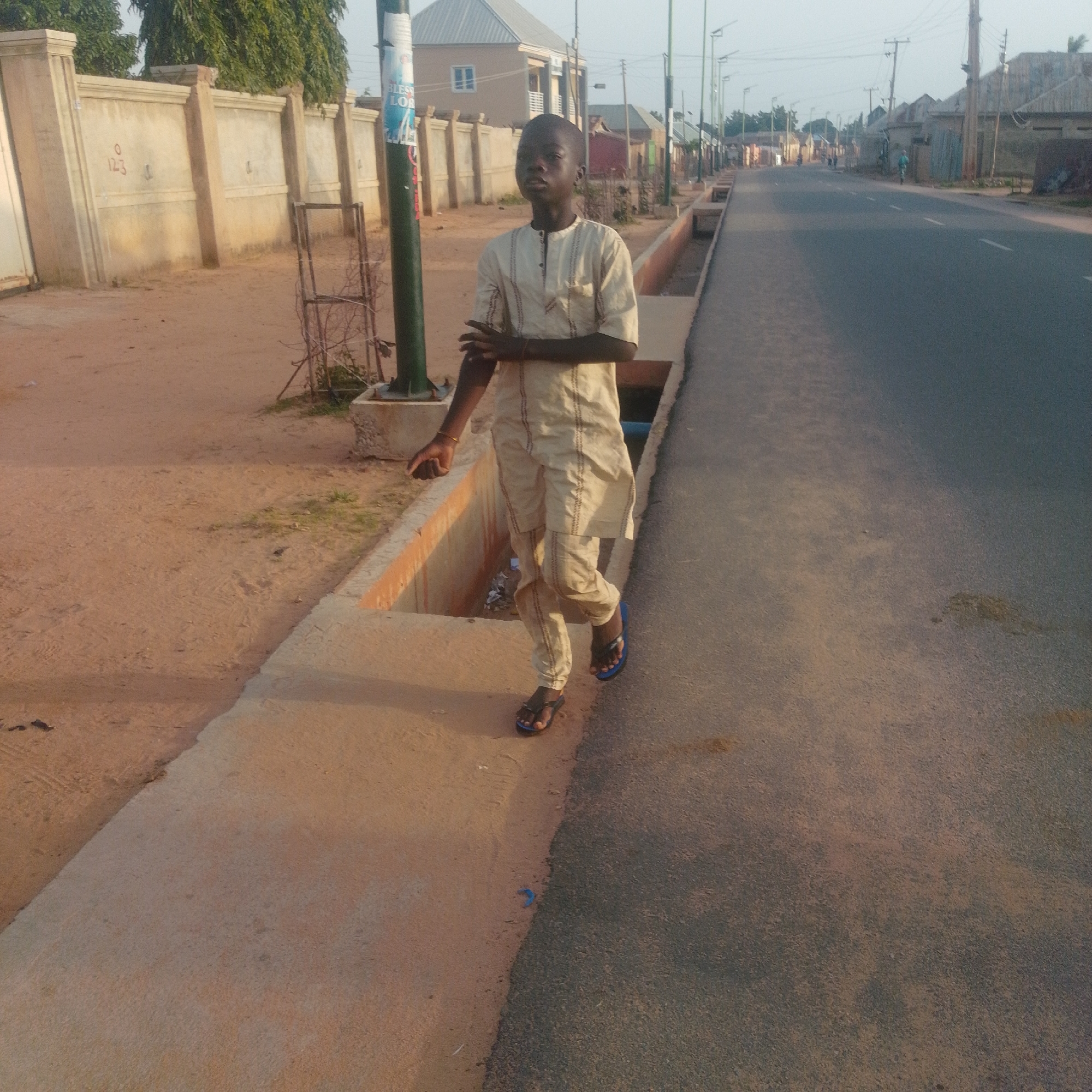 A young Black male, wearing a light-colored traditional outfit and blue flip-flops, walks along a concrete path next to a paved road. The boy is walking towards the right of the frame with his left hand outstretched, palm open, and his right arm bent at the elbow. Behind him to the left is a sandy area with a beige concrete wall and some trees. To the right of the boy is a street with streetlights and buildings in the background. The sky is overcast.