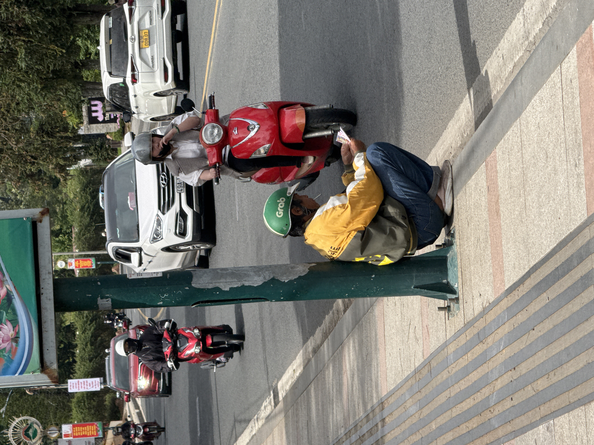 On a paved city street in Da Lat, Vietnam, a person wearing a green and yellow jacket, blue jeans, and a green helmet crouches by a dark green utility pole. They are holding some papers in their hands and appear to be smoking a cigarette, with the lit end visible. In the background, several motorbikes and cars are driving on the road. One red scooter is close to the crouching person, and another red scooter is further up the road carrying a person with a white shirt and vest. A white SUV is also visible in the mid-ground. The sidewalk beside the road has raised tactile paving. The sky is overcast.