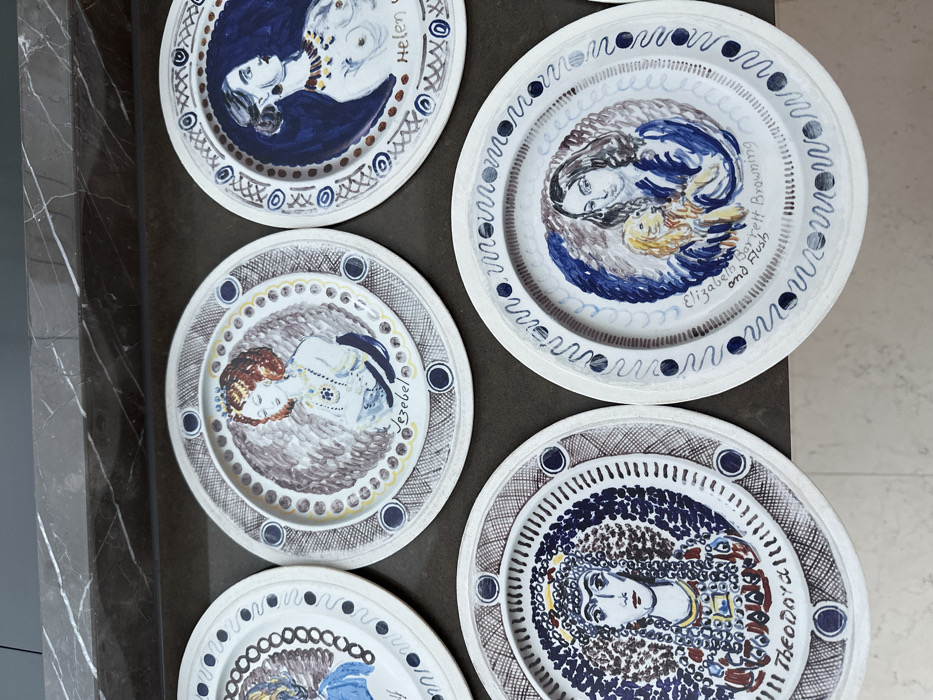 The image shows a collection of decorative plates with hand-painted designs. Each plate features a portrait within a circular frame. Some plates depict women, such as Helen, Jezebel, and Theodora, accompanied by their names. Another plate illustrates Elizabeth Barrett Browning with her dog, Flush. The plates have a folk art style, characterized by simple lines and bold colors. The borders include geometric patterns and repetitive motifs like dots, lines, and circles.