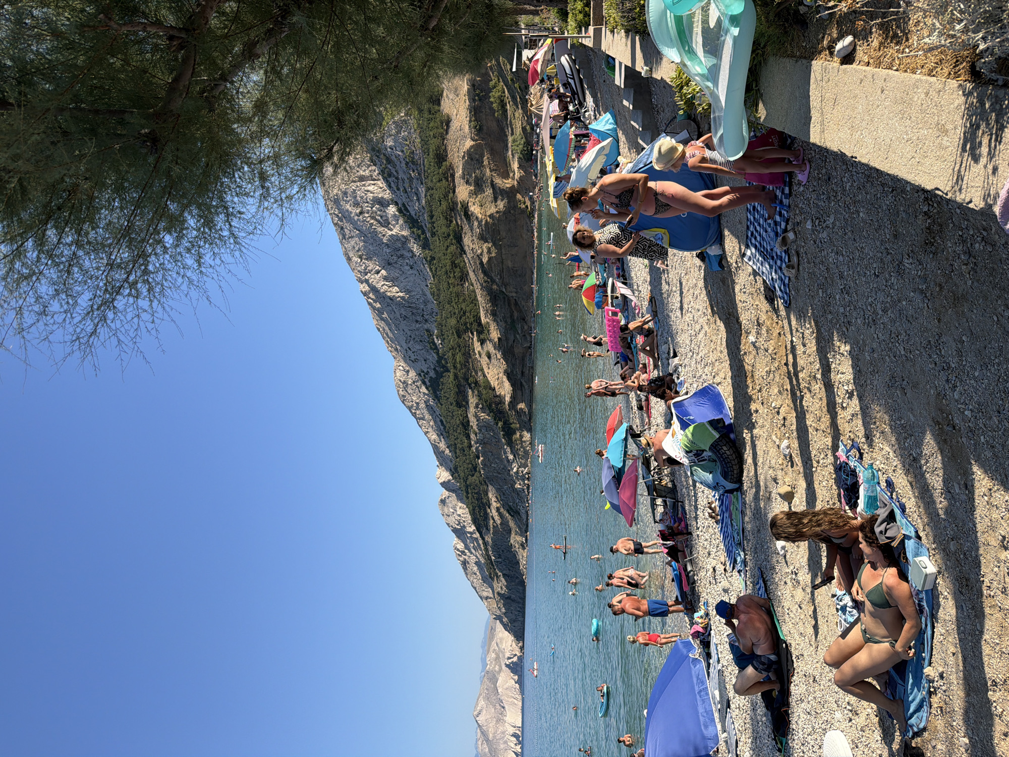The image depicts a crowded pebble beach in Baška, Općina Baška, Croatia, under a clear blue sky. Numerous people are engaged in various beach activities: swimming in the clear turquoise water, sunbathing on towels and loungers, and standing near umbrellas. Several colorful beach umbrellas provide shade. The background features a rugged, rocky mountainside dotted with sparse green vegetation, sloping down towards the sea. The water is calm, with a few small boats visible in the distance. The shoreline is lined with people, some interacting, while others are relaxing individually. The overall scene conveys a typical summer day at a popular coastal resort.