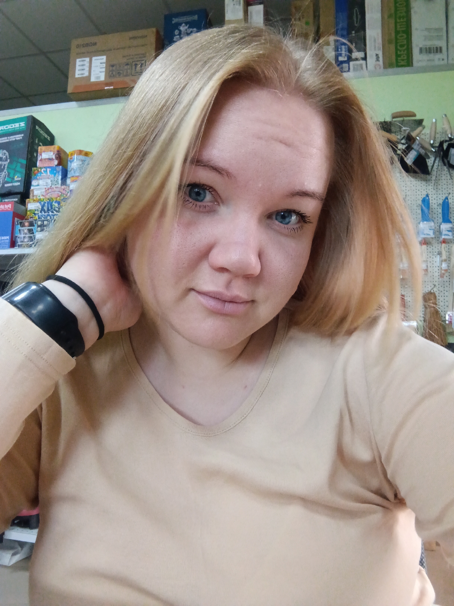 A woman with blonde hair and blue eyes is taking a selfie in a store. She is wearing a beige t-shirt and a black bracelet on her left wrist. The background shows shelves stocked with various items, including boxes and some tools. The location is identified as a store in the city of Fokino, Russia.