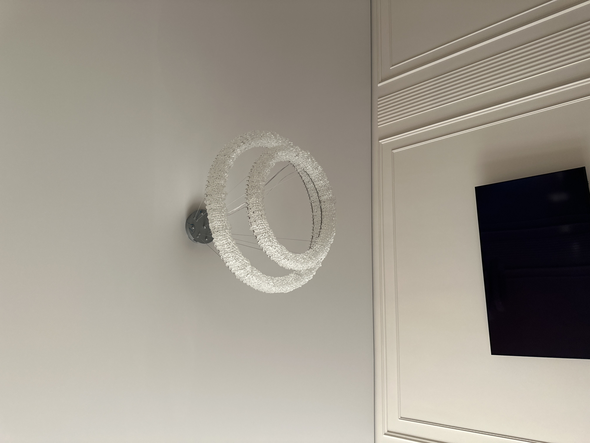 A modern crystal chandelier hangs from a white ceiling in a room. The chandelier consists of two intertwined rings adorned with numerous clear crystals. Below the chandelier, a white wall features decorative molding, creating a wainscoting effect. A portion of a dark, flat-screen television is visible mounted on the wall. The scene appears to be indoors, in a residential or commercial space. No specific location in Almaty, Kazakhstan can be identified from the visual information provided.