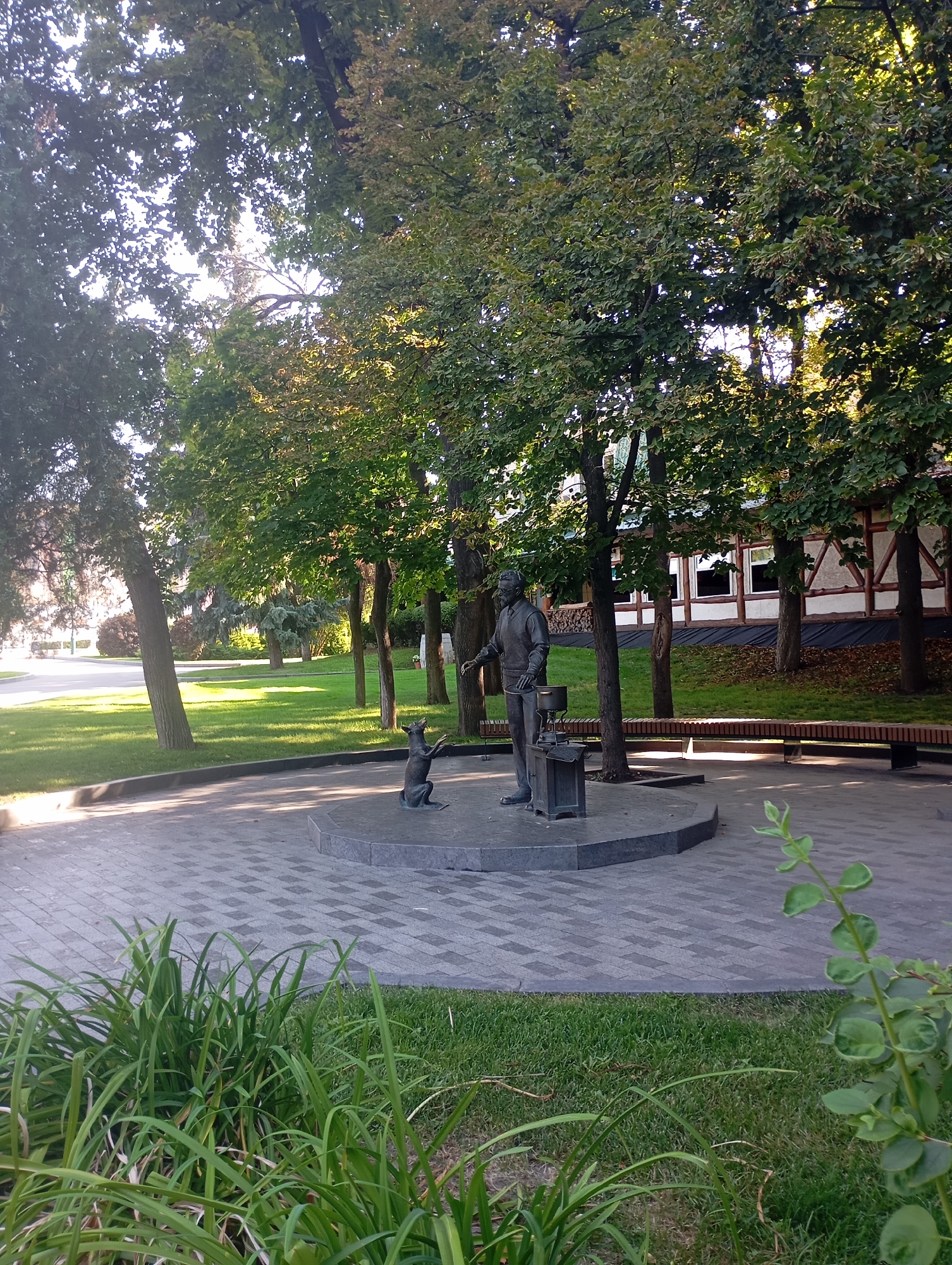 This is a full-shot, eye-level, outdoor photograph of a bronze sculpture of a man and a dog in a park setting. The man, depicted in a suit and tie, is shown reaching down towards the dog, which is on its hind legs, appearing to interact with the man. The sculpture stands on a circular, multi-tiered stone pedestal. To the right of the pedestal, a long, wooden bench is visible.
The park features lush green grass and mature trees with dense foliage, casting shadows on the paved walkway. In the background, a building with a timbered facade and large windows is visible. The scene is illuminated by natural daylight.
This sculpture is located in Kharkiv, Ukraine, specifically in Kharkiv City Park, a well-known public space. It is a monument dedicated to the famous Soviet writer Mikhail Bulgakov and his dog, Flay. The sculpture depicts Bulgakov interacting with his beloved pet, and it has become an iconic symbol of the city.