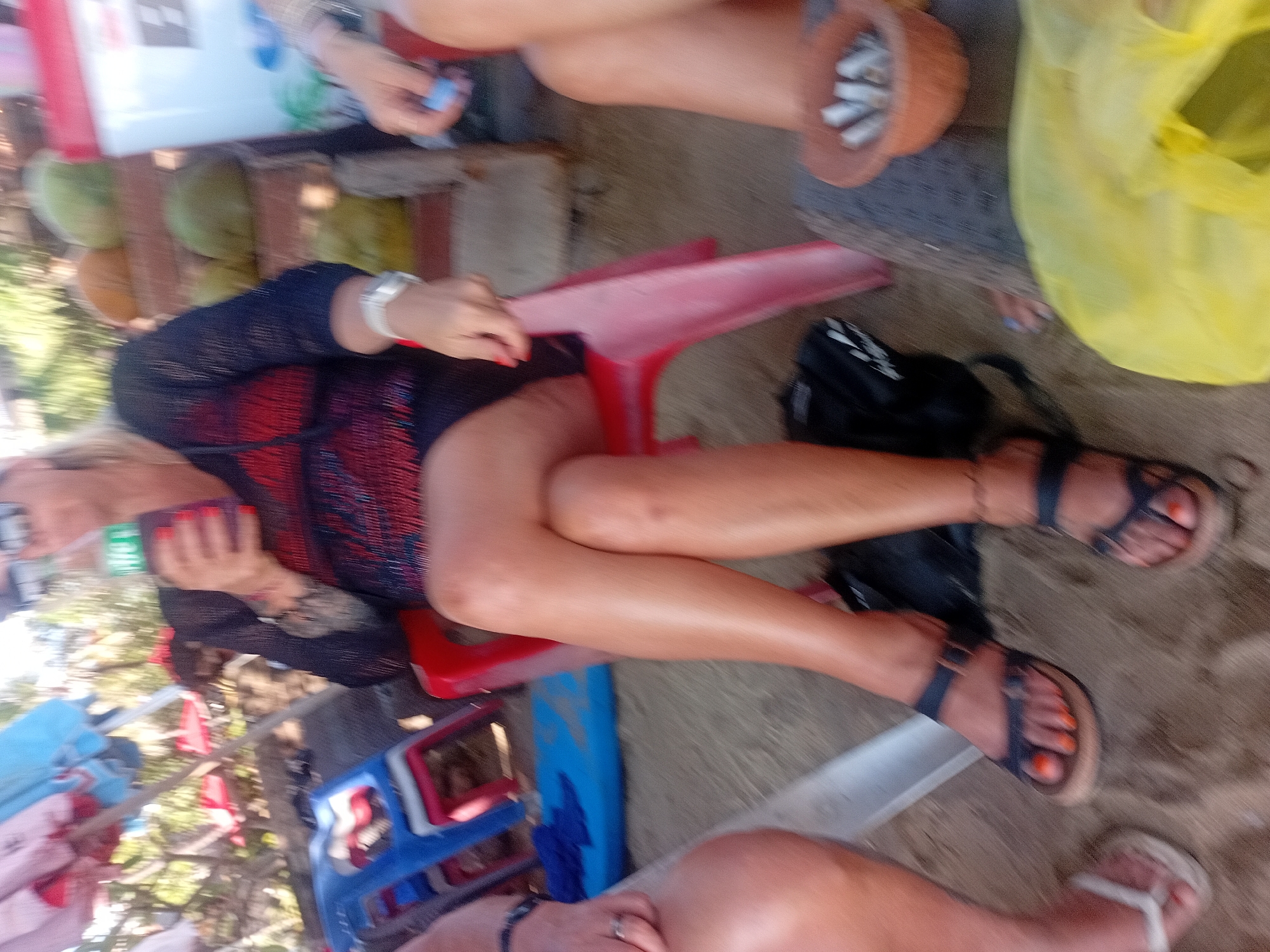 The image shows three individuals sitting on chairs on a sandy surface, likely a beach in Kuta, Indonesia. In the foreground, a person is wearing dark sandals with orange painted toenails and a black anklet. Beside them, another person's legs are visible, with one foot wearing a white flip-flop. To the right, a third person is partially visible, their legs extending towards the foreground, and they appear to be holding a light blue object.
The person in the center is wearing a black, sheer, long-sleeved top over a red and black patterned garment. They are holding a bottle with a green label. Their legs are crossed at the knees. Behind them, red and blue plastic chairs are stacked, and in the background, there are indications of beach umbrellas and trees. On the far right, a small brown clay pot filled with cigarette butts is visible, resting on what appears to be a woven mat. A yellow plastic bag is also present in the lower right quadrant of the image. The overall lighting suggests it is daytime, likely outdoors.