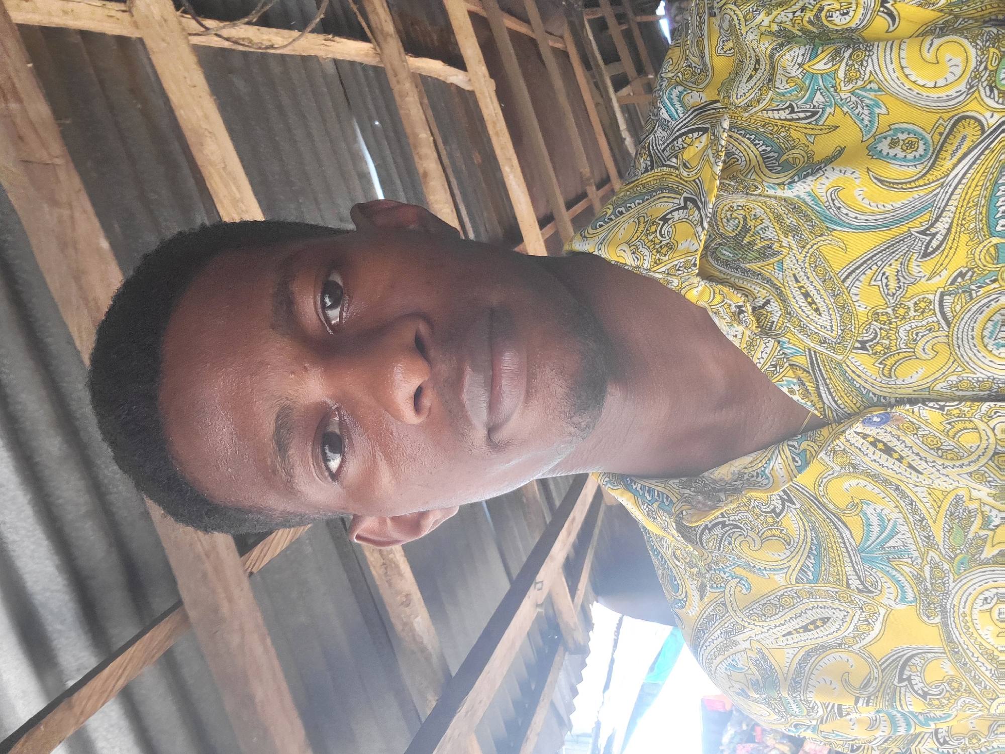 A close-up, head-and-shoulders portrait of a young Black man facing forward, captured in an outdoor setting, likely a market or covered stall in Onitsha, Nigeria, due to the corrugated metal roofing and visible wooden beams. The man has short, dark hair, dark eyes, and a light stubble. He is wearing a bright yellow shirt with a paisley pattern in shades of blue, green, and white. The lighting suggests it is daytime, with sunlight creating some glare on his face and shirt. In the background, the textured ceiling of the structure is visible, along with some faint shapes of other people or merchandise, but they are out of focus and do not provide specific identifying details. The overall impression is a casual, candid photograph taken in a bustling environment.