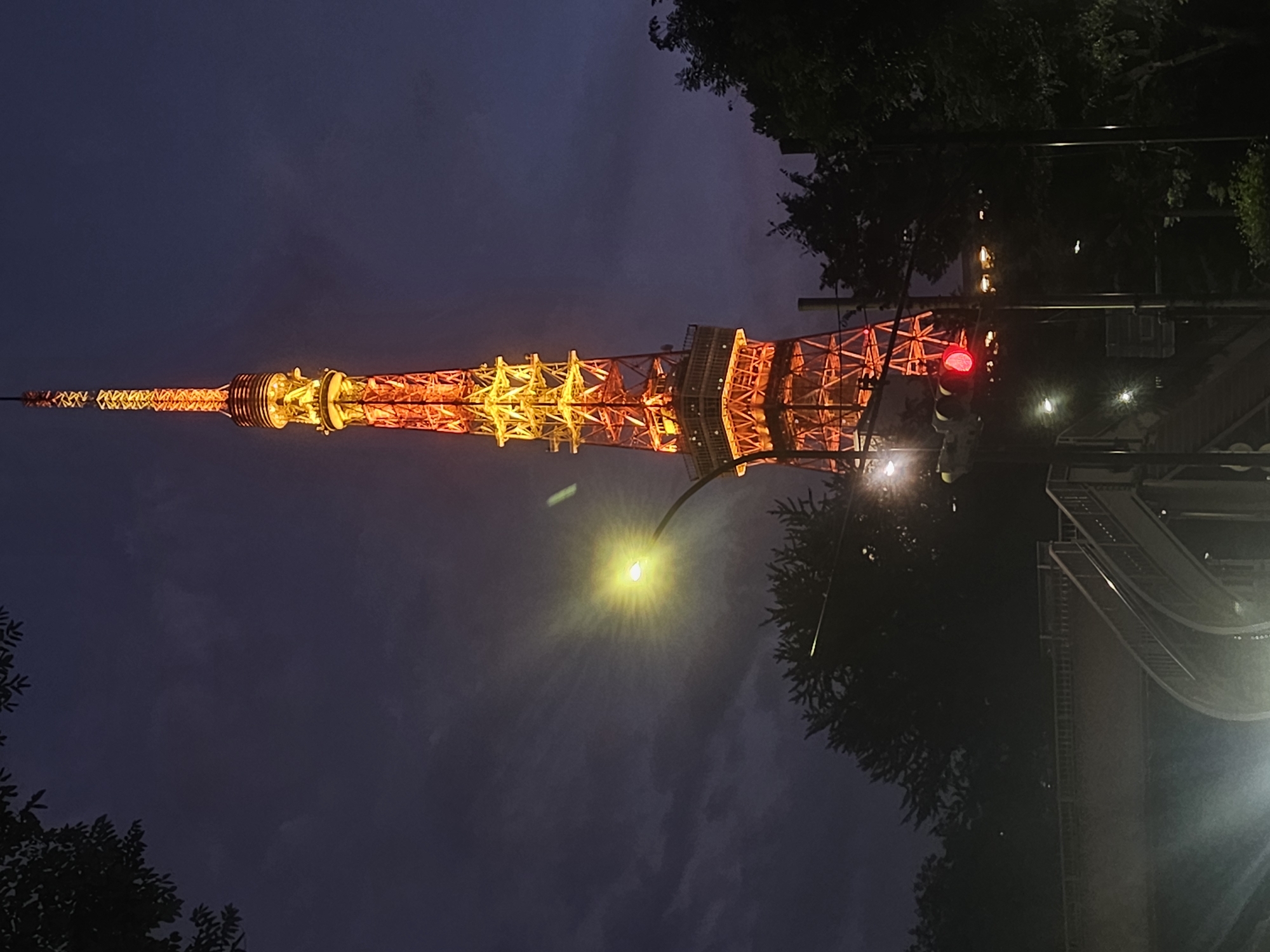 The image captures the iconic Tokyo Tower in Minato, Japan, illuminated with a vibrant orange and yellow glow against a dark, cloudy twilight sky. The tower, a prominent landmark, is the central subject, its intricate lattice structure clearly visible. In the foreground, a street light emits a bright, starburst-like glow, casting a yellow hue. Adjacent to it, a traffic light displays a vivid red signal. A pedestrian overpass with railings and stairs is visible in the lower left portion of the frame, partially obscured by darkness and the overhanging branches of trees that frame the scene on the left and right. The overall impression is one of urban nightfall, with the illuminated tower dominating the composition.