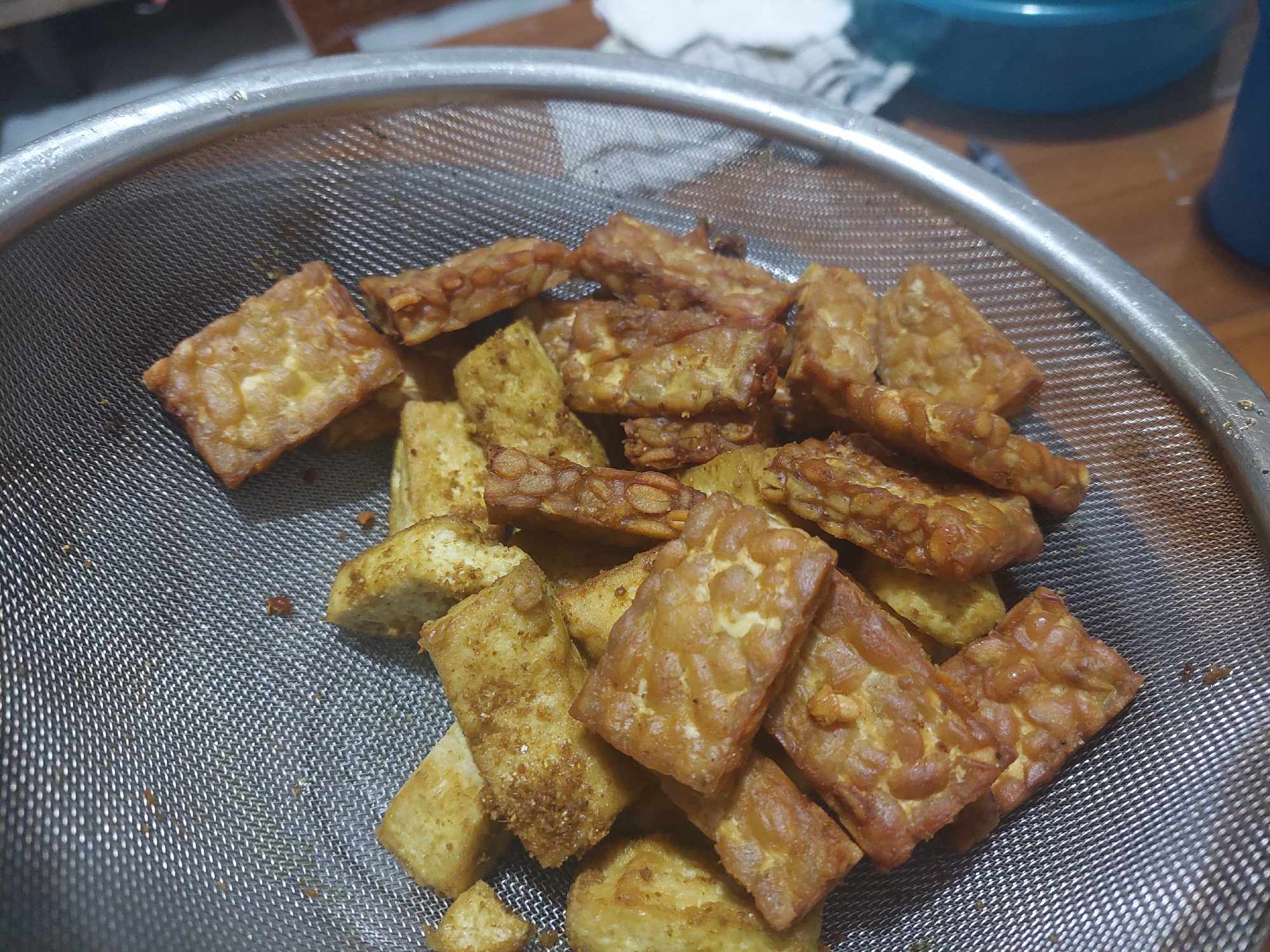A close-up, overhead shot shows a pile of fried tempeh and tofu in a metal colander. The tempeh pieces are rectangular and have a visible grain pattern, while the tofu pieces are irregular in shape. Both are golden brown and appear to be slightly greasy. The colander is placed on a wooden surface, and in the background, a blue bowl and a partially visible blue container are present. The scene is indoors and appears to be a kitchen setting. The image was captured in Jelutung, Indonesia.