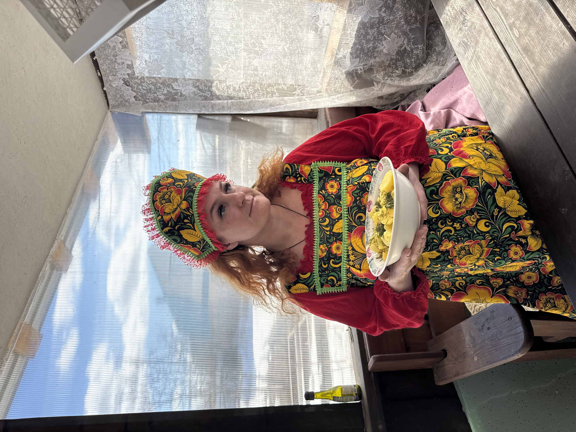 A woman in traditional Russian clothing holds a bowl of food inside a greenhouse or sunroom, likely in Chaikovsky, Russia. The woman wears a red blouse with a floral patterned dress and matching headdress. She is holding a white bowl filled with what appears to be boiled potatoes, garnished with dill. The background consists of translucent corrugated plastic panels of the greenhouse walls, with a white lace curtain visible above. A wooden bench is visible on the right side of the frame, and a bottle sits on the floor near the corner of the wall.