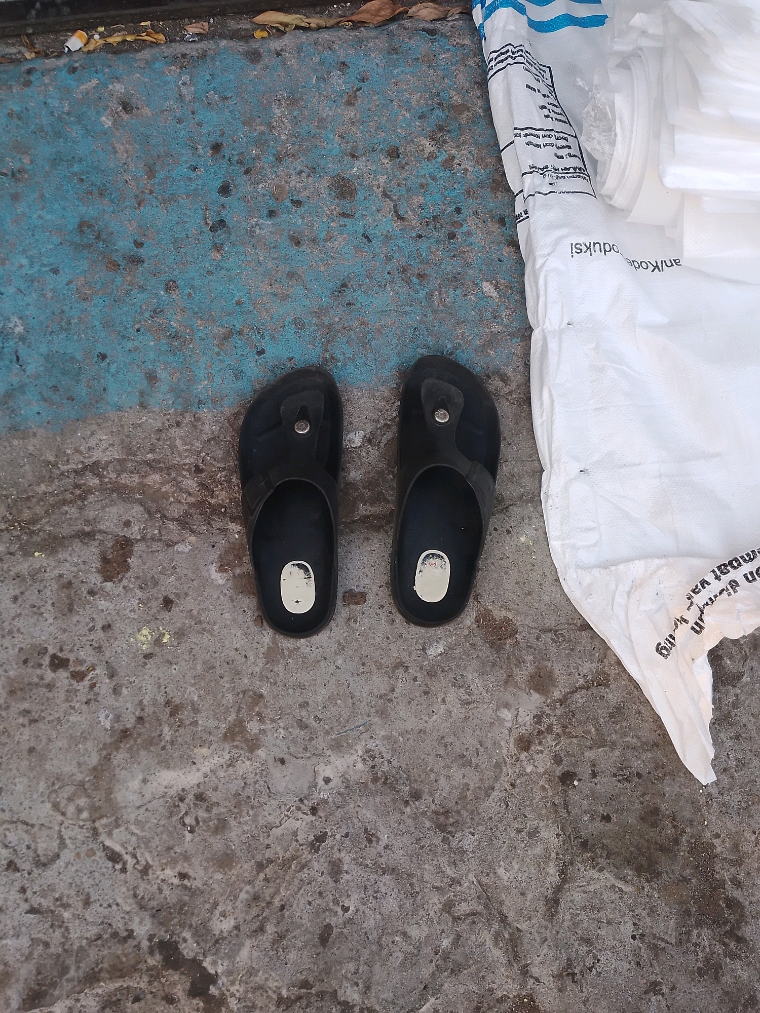 A pair of black flip-flops are situated on a concrete surface, adjacent to a large white plastic bag. The concrete is partially covered by a faded blue painted area. In the background, a portion of the white plastic bag is visible, bearing text and blue stripes. The overall environment suggests an outdoor setting, possibly a roadside or public area in Patianrowo, Indonesia. There are no people or animals present in the image. The scene is static and appears undisturbed.