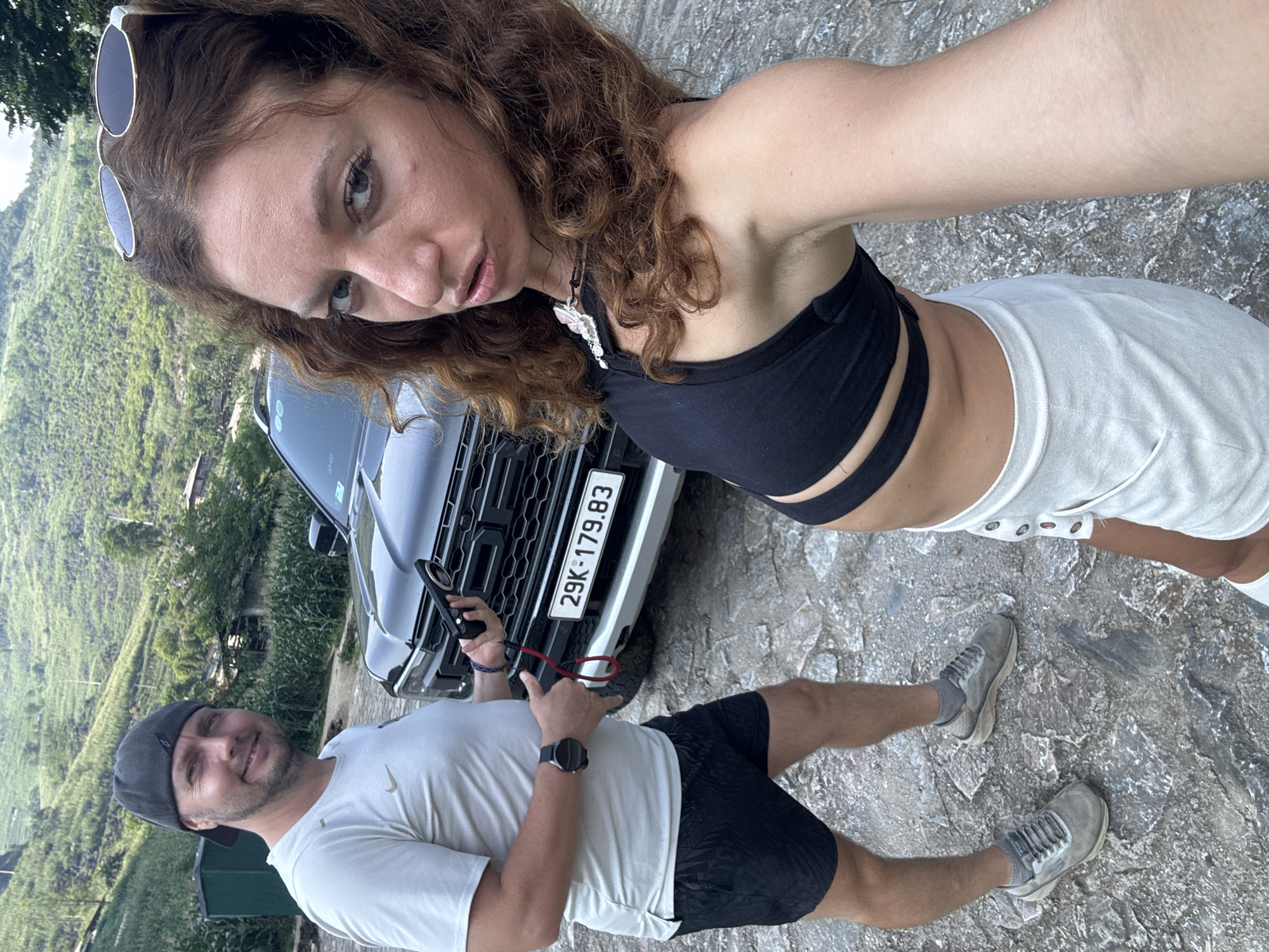 A woman and a man pose for a photo near a gray car with license plate 29K-179.83. The woman wears sunglasses on her head, a black top, and white shorts. The man wears a cap, a white t-shirt, and black shorts, holding a camera. The background shows a landscape of Đường Thượng Commune, Vietnam. The ground is a stony surface.