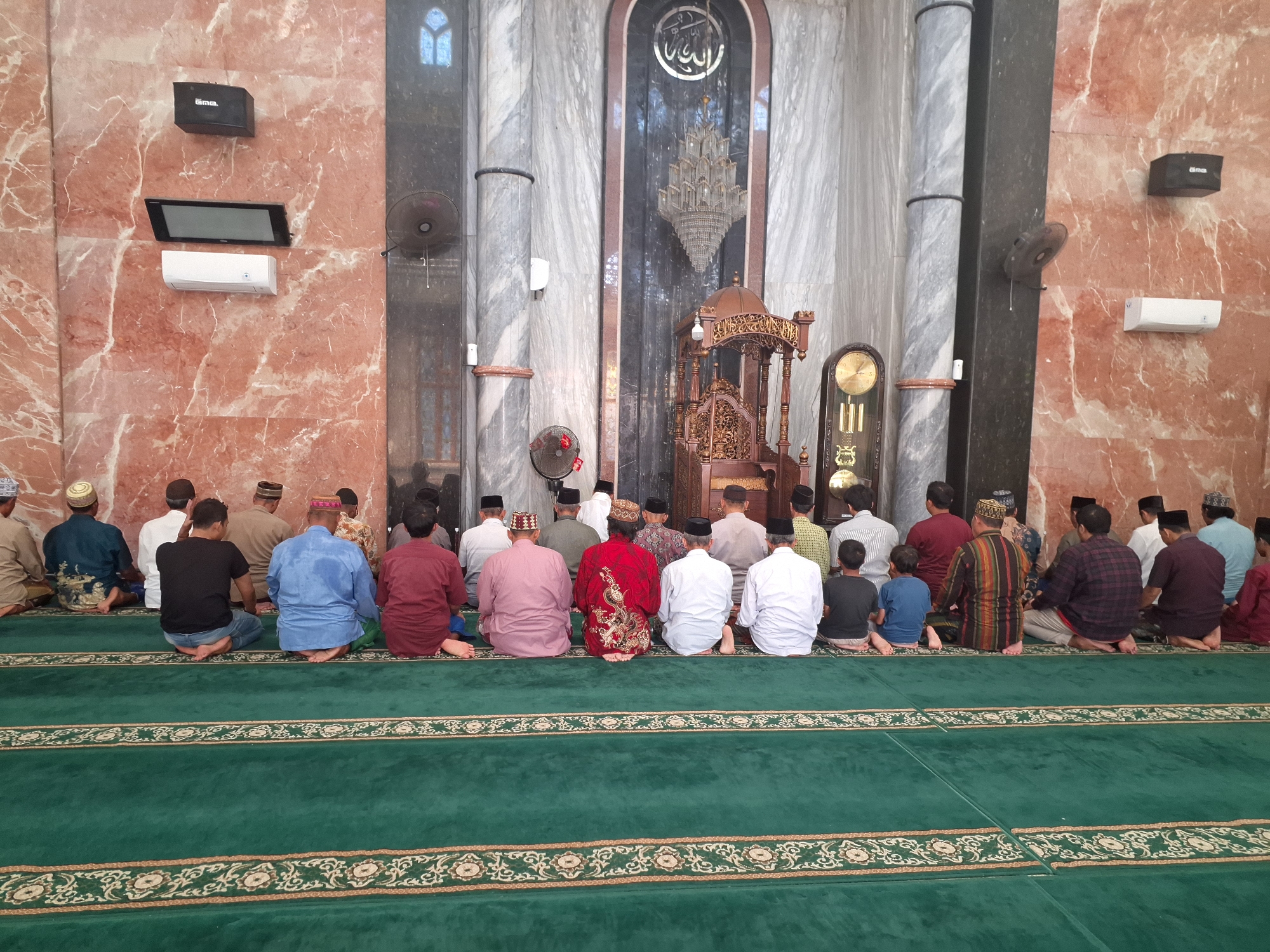 The image depicts the interior of a mosque in Surabaya, Indonesia, with a group of men and boys engaged in prayer. The men are seated on a green carpet with intricate patterns, facing the mihrab. Their attire varies, with some wearing traditional religious clothing and others in more casual attire. The mosque's interior features marble walls, ornate pillars, a large chandelier, and a wooden pulpit. In the background, a grandfather clock and fans are visible. The overall scene conveys a sense of solemnity and devotion.