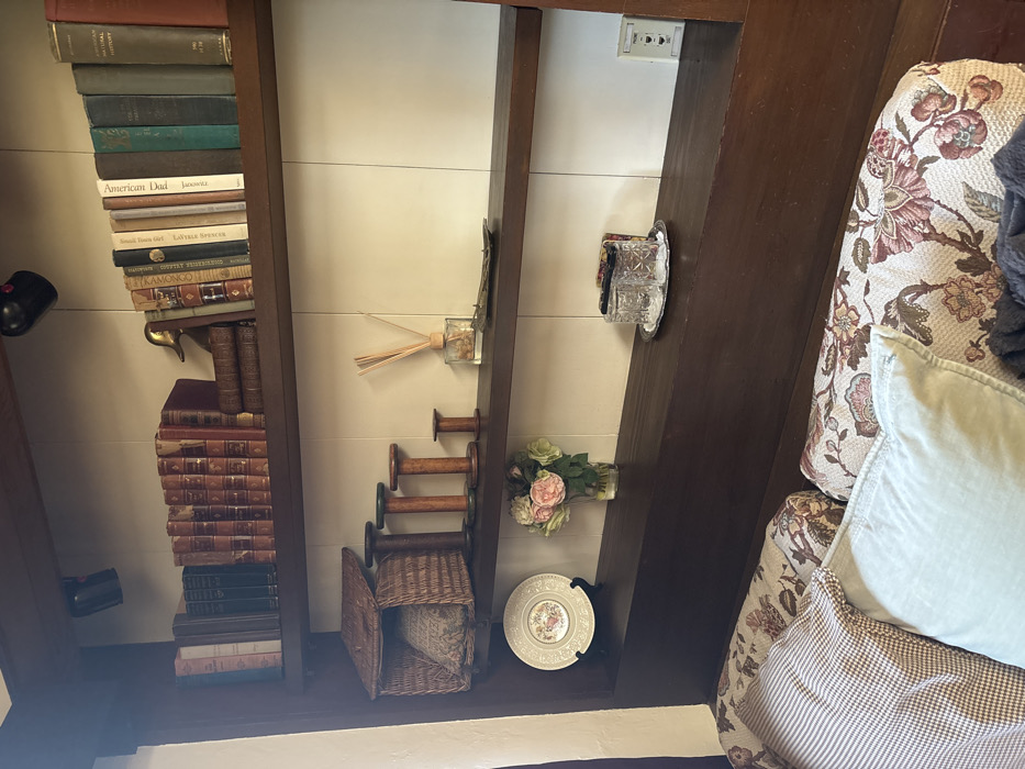 This interior scene depicts a dark wood built-in shelving unit above a seating area, located in Monterey, United States of America.
The top shelf contains approximately 30 books, varying in size and color. A collection of reddish-brown bound books is centrally located, flanked by other books with dark and light-colored spines, one titled "American Natural History." A gold-colored duck figurine stands on the right, functioning as a bookend, with two dark-spined books stacked horizontally beneath it. Two black wall-mounted spotlights are visible above this shelf.
The middle shelf features a wicker basket with a hinged lid, containing a patterned cushion. To its right are four wooden spools or bobbins, graduated in height, with the tallest exhibiting a green-painted top edge. Further right, a clear glass reed diffuser bottle with reeds and a small, dark, ornate tray are present.
The bottom shelf displays a decorative plate with a floral pattern, propped vertically on a dark stand. Next to it, a clear glass vase holds a bouquet of artificial pink and green roses. On the right, a silver-colored oval tray contains two clear, patterned drinking glasses and a stack of dark coasters. A white wall-mounted panel with "PHONE" and "TV" labels and three small icons is visible on the far right.
Below the shelving unit, the top portion of a seating arrangement, likely a sofa or bench, is visible. It is upholstered in a floral pattern featuring pink, green, and brown tones. Multiple throw pillows are present, including one with a light brown houndstooth pattern and another in a solid light blue color. A dark fabric is partially visible beneath the pillows. The wall behind the shelves is white with vertical paneling. No individuals are depicted.