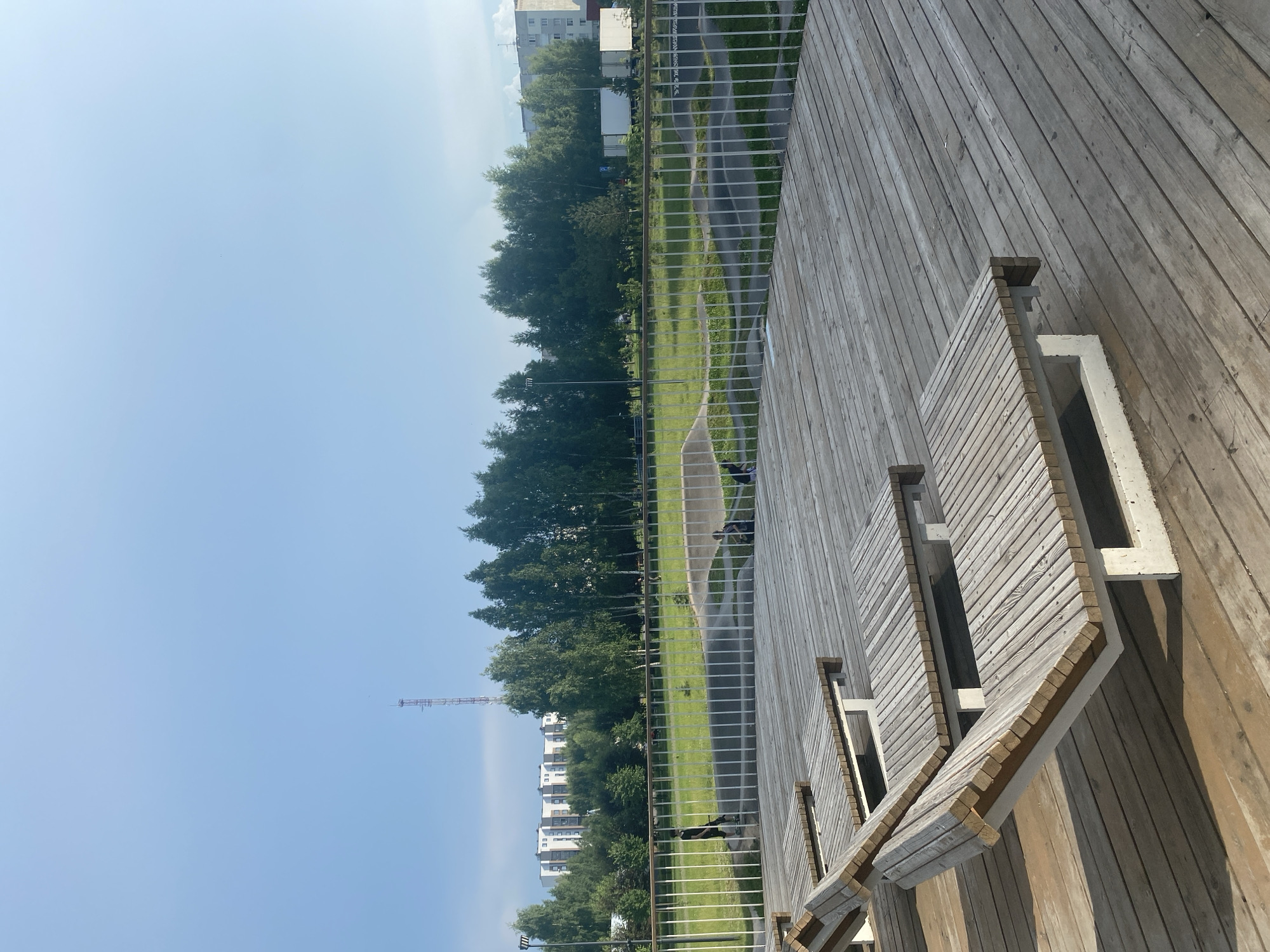 The image, taken in Zelenodol'sk, Russia, captures a portion of a modern outdoor public space with a wooden deck and seating. The scene is oriented vertically, with the sky occupying the top left portion of the frame and a wooden deck filling the right side.
The deck is made of weathered wooden planks, arranged diagonally from top right to bottom left. Integrated into the deck are several built-in benches, constructed from lighter-colored wooden slats with white concrete supports. These benches are designed with angled seating and some feature empty rectangular recesses.
Beyond the deck, a metal railing with vertical bars separates the elevated area from a grassy park. The park features a paved pathway winding through it, and is lined with a dense row of mature green trees. In the background, several multi-story apartment buildings are visible, interspersed with more trees. A tall, lattice-like tower, possibly a radio or cell tower, stands to the left of the buildings.
The sky is a clear, pale blue, suggesting a sunny day, likely in the late morning or early afternoon due to the shadows cast on the deck. A few dark specks in the sky might be birds. The overall atmosphere is calm and open, typical of a park or public recreational area. No specific activities are clearly depicted, though the presence of benches suggests a place for rest or observation. There is no visible text in the image.