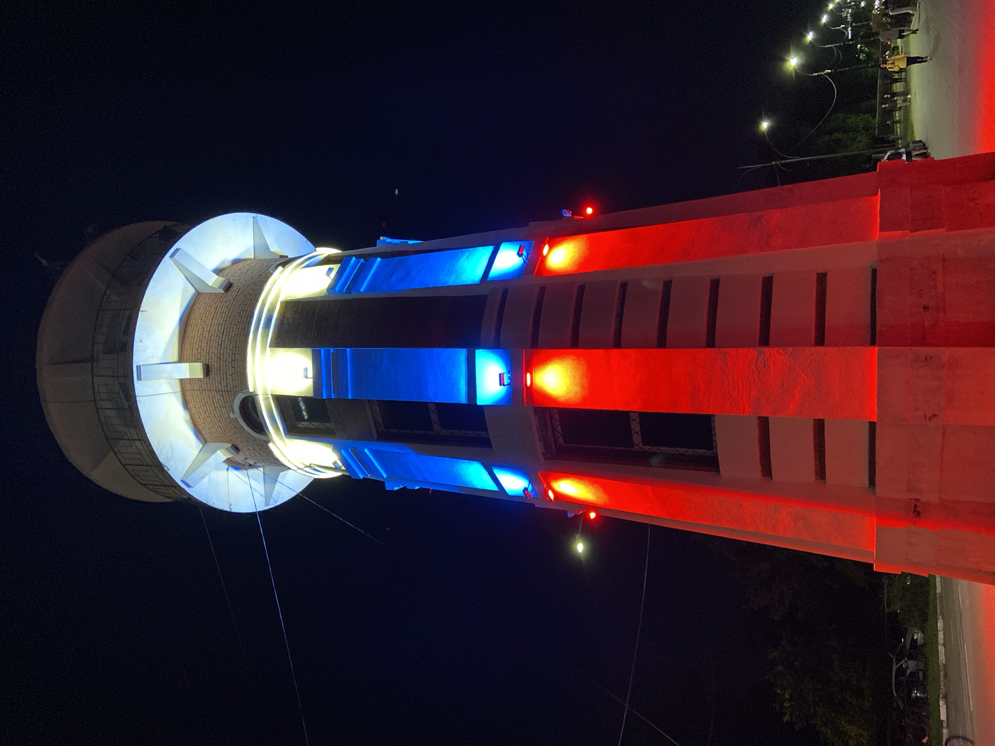 This is a night-time, low-angle, full-frame shot of the Blagoveshchensk Water Tower, located in Blagoveshchensk, Russia. The tower is illuminated with red and blue lights, creating a striking visual against the dark night sky. The upper portion of the tower is capped with a round, white structure with a railing, and is lit with white and yellow lights. Below this, the brick structure of the tower is illuminated with vertical bands of blue and red light. The base of the tower is predominantly red. In the background, to the right, a street with lampposts and a sidewalk is visible, with several people walking. Trees and parked cars are visible on the left side of the image.