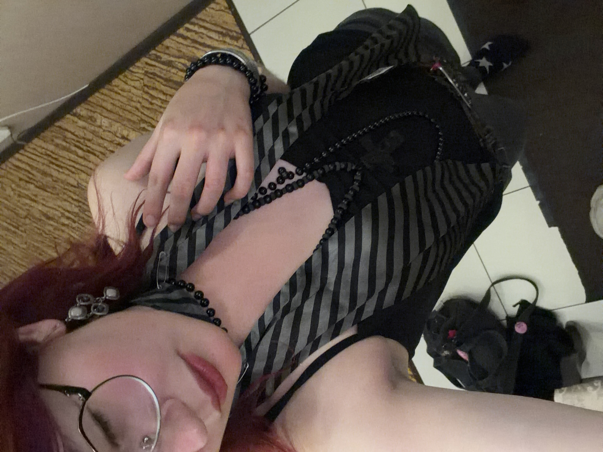 A person with dark red hair and glasses is taking a selfie. They are wearing a black tank top with a striped scarf draped around their neck and layered necklaces. They also have a beaded bracelet on their left wrist and decorative earrings. On the floor, there is a black garment, possibly a bra, and a sock with white stars. The background appears to be a carpeted surface. The image does not contain any identifiable landmarks or context to place it in Chelyabinsk, Russia.