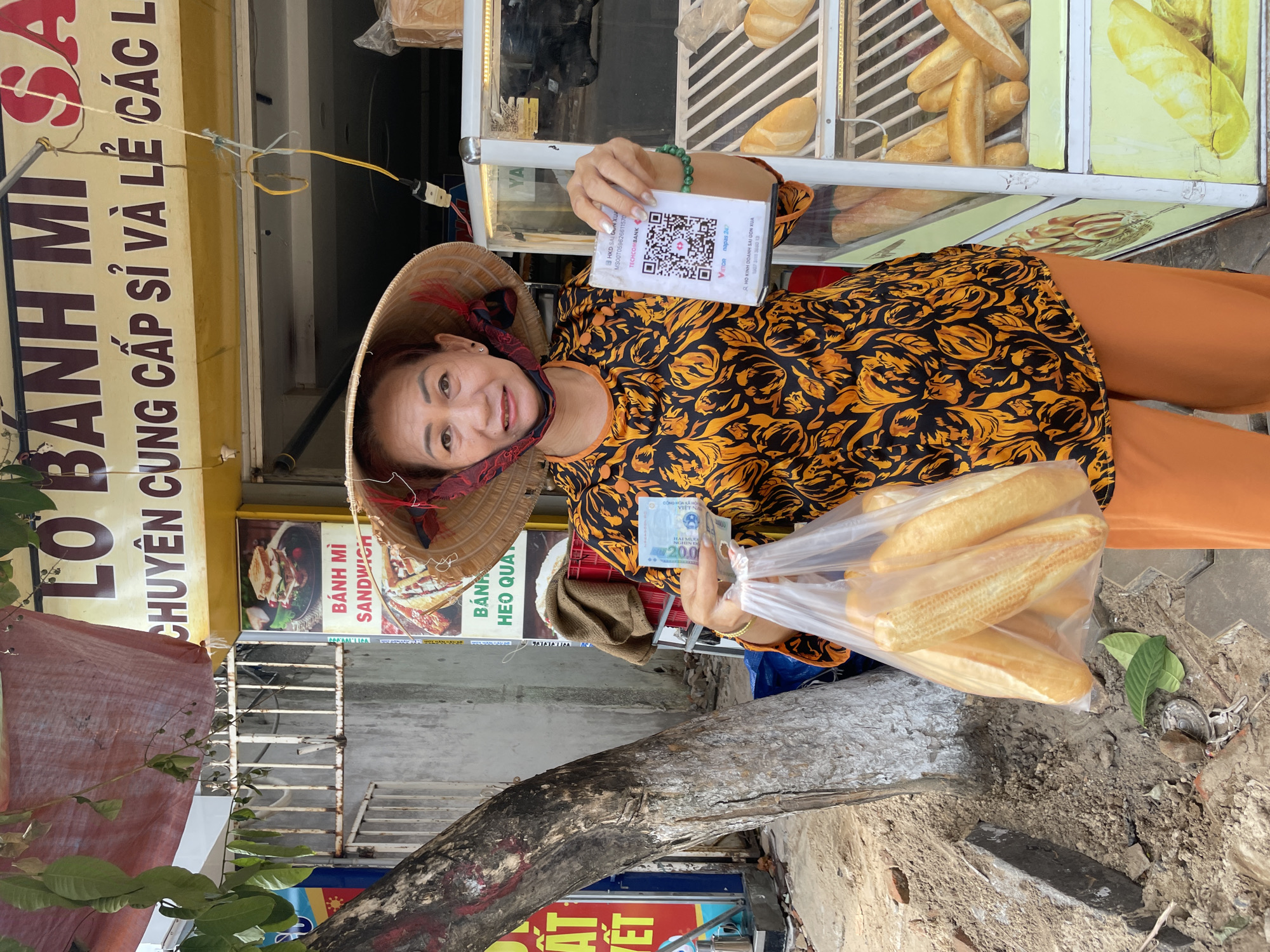 A middle-aged Asian woman, wearing a conical hat and a vibrant orange and black patterned shirt, stands on a sandy and paved street in Phu Thuy Ward, Vietnam. She is holding a plastic bag filled with freshly baked baguettes in her left hand and a Vietnamese 200,000 dong bill and a payment QR code in her right. The QR code is for Techcombank and displays logos for "Vnnpay" and "napaps 24/7". Behind her, a food stall displays more baguettes, and signage indicates it sells "Banh Mi". The scene appears to be a typical street-side vendor operation.