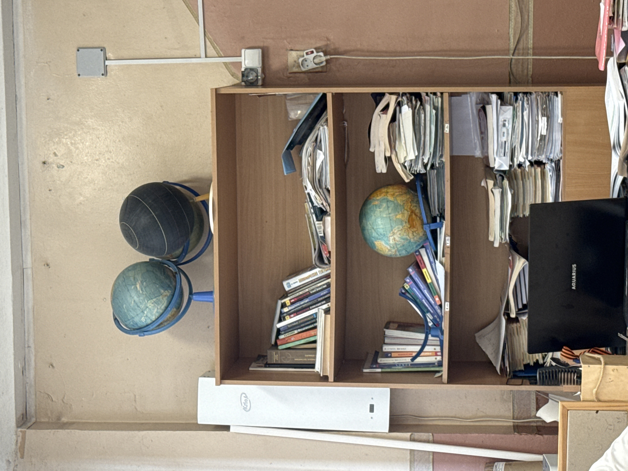 The image depicts a cluttered shelf in what appears to be an indoor setting. The shelves are made of light-colored wood. Two globes, one black and one blue, are positioned to the left of the shelves. Inside the shelves are piles of books, papers, and hanging folders. A black binder with the word "AQUARIUS" on it is visible on the right side of the shelves. A white box with the inscription "Ictio" is situated under the shelves. The walls are painted in a pale peach color, and there is evidence of peeling paint and wear. An electrical outlet and a light fixture are visible near the top of the frame. The image does not provide enough information to determine the specific location in Saint Petersburg, Russia, or any details about people or actions.