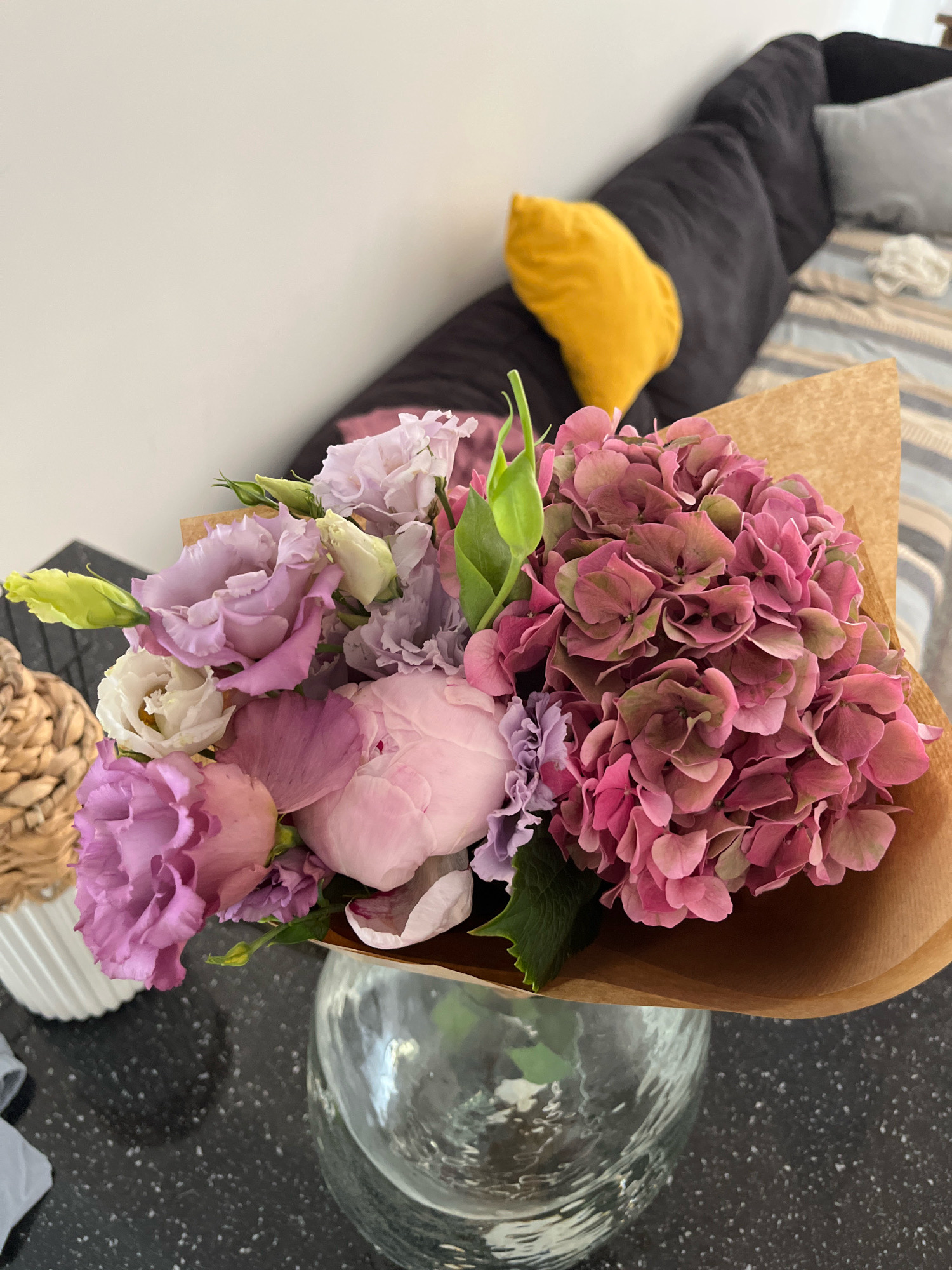 An indoor still life scene features a bouquet of pink and purple flowers, primarily hydrangeas and roses, arranged in a clear glass vase. The flowers are partially wrapped in brown paper. The bouquet is placed on a dark, speckled countertop. In the background, a dark sofa with a yellow pillow is visible, along with a striped cushion on the sofa and a white decorative object to the left of the flowers. The scene is well-lit. The location is Limassol, Cyprus.