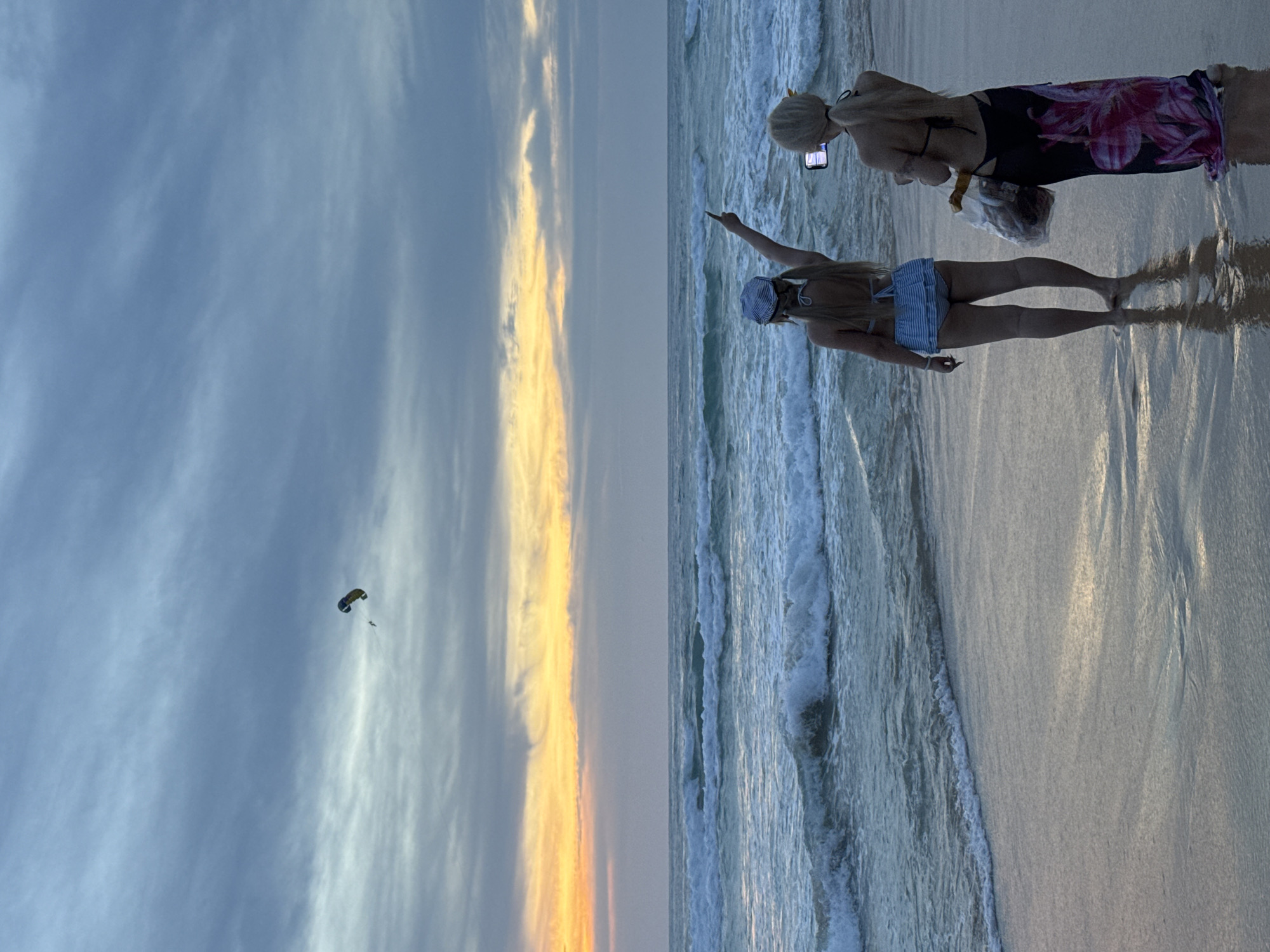 Two women are standing on Karon Beach, Thailand, facing the ocean. The sky is overcast with streaks of orange and yellow from the setting sun. The woman on the left, wearing a blue and white striped bikini and a cap, is pointing towards the sky where a paraglider is visible. The woman on the right is holding a smartphone and taking a picture. She is wearing a black bikini and a colorful sarong. The ocean has moderate waves, and the wet sand reflects the twilight sky.