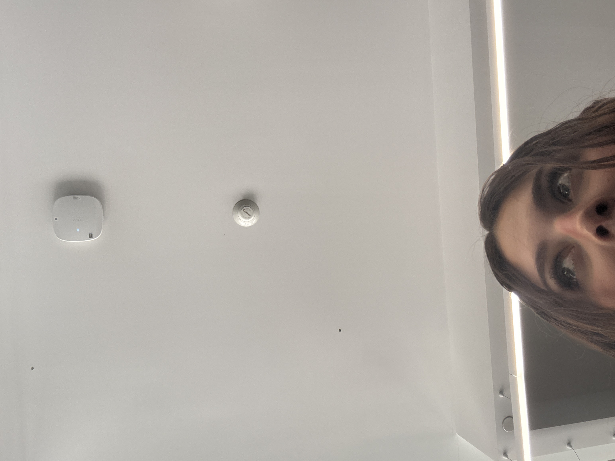 The image provides an upward-angle view within an indoor environment, primarily featuring a white ceiling and the partial upper facial area of an individual. The white ceiling displays a rectangular, rounded white device with a faint blue indicator light, positioned towards the upper right. Below this, a smaller, circular white smoke detector is mounted. A bright, horizontal linear light fixture, possibly integrated into a wall-ceiling junction, is visible along the lower left edge of the ceiling. Several small, white, circular protrusions are also present on the ceiling in the lower left quadrant. Two minor, dark circular marks are visible on the plain ceiling surface. The bottom portion of the frame captures the forehead, eyes, and upper nose of a person with dark brown hair, whose gaze is directed upwards and to the right. The specific location within Mexico City, Mexico, is not discernible from the visual evidence.