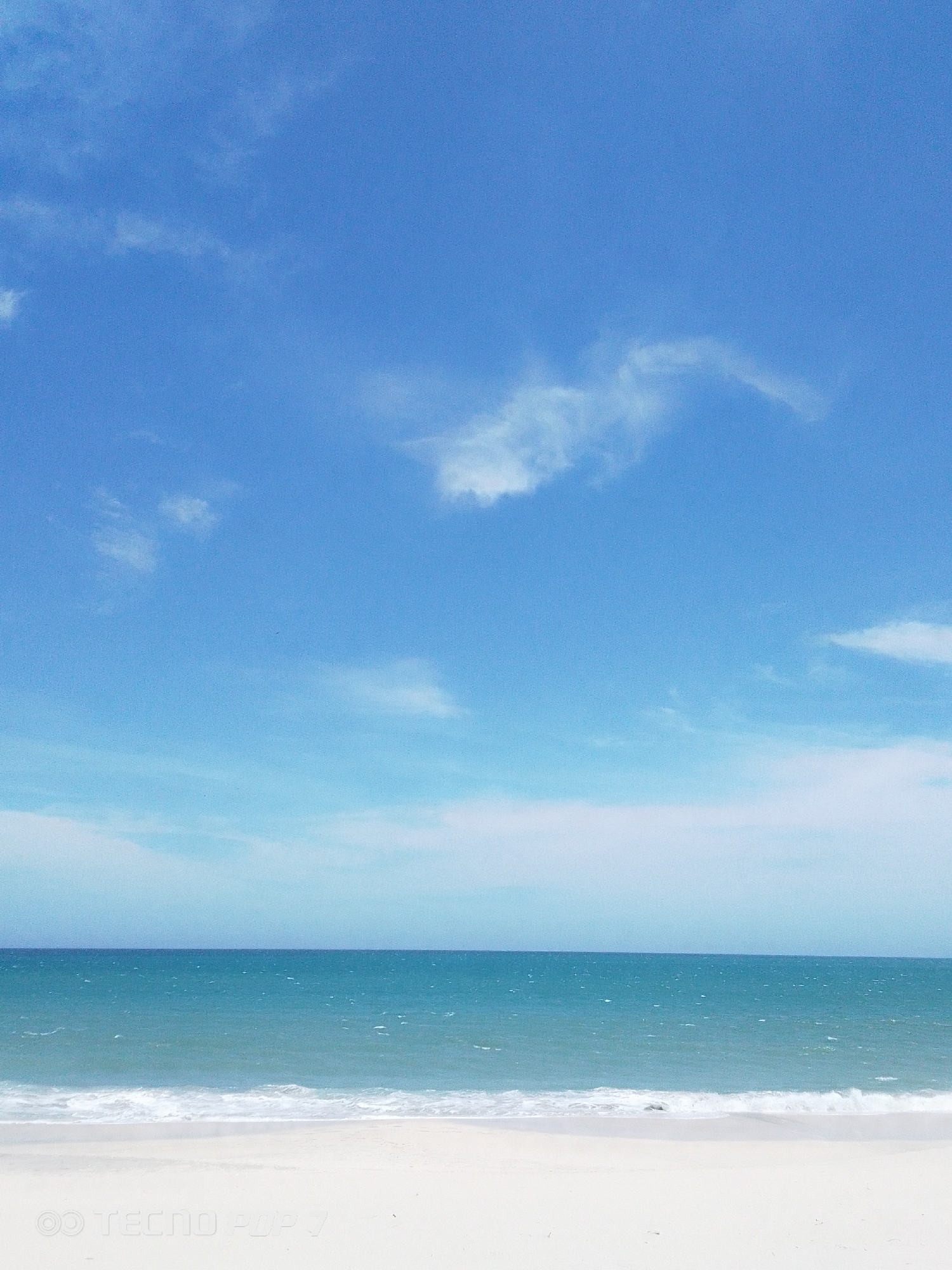 The image depicts a coastal scene, likely a beach in Pedro González, Venezuela. The sky is a bright blue with scattered white clouds. The sea is a turquoise-blue with small waves breaking near the white sandy beach. No people are visible in the scene. The photograph appears to have been taken on a clear, sunny day. In the bottom left corner, a watermark is visible "TECNO POP V."
