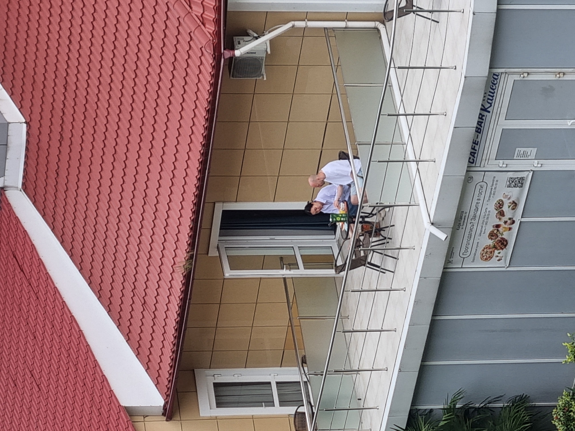 Here is a description of the image.
The image is an upward, angled, outdoor shot of a building, possibly in the city of Sochi, Russia. The building is multiple stories, with light brown tile paneling on the walls. The roof is red with a metal shingle design. There is a white border along the roof’s edge.
There are two men sitting on a balcony on the upper level of the building. They are both wearing white shirts. One man appears to be balding. The men are seated at a table with some objects on it, possibly drinks or food. There are metal railings that frame the balcony.
To the right of the balcony is a vertical sign that reads "CAFE-BAR Kaйсса". Below the sign is a poster with images of food and Russian text. To the right of the building, at the bottom of the image, are the tops of some green plants.
The image is well-lit, suggesting it was taken during the day.