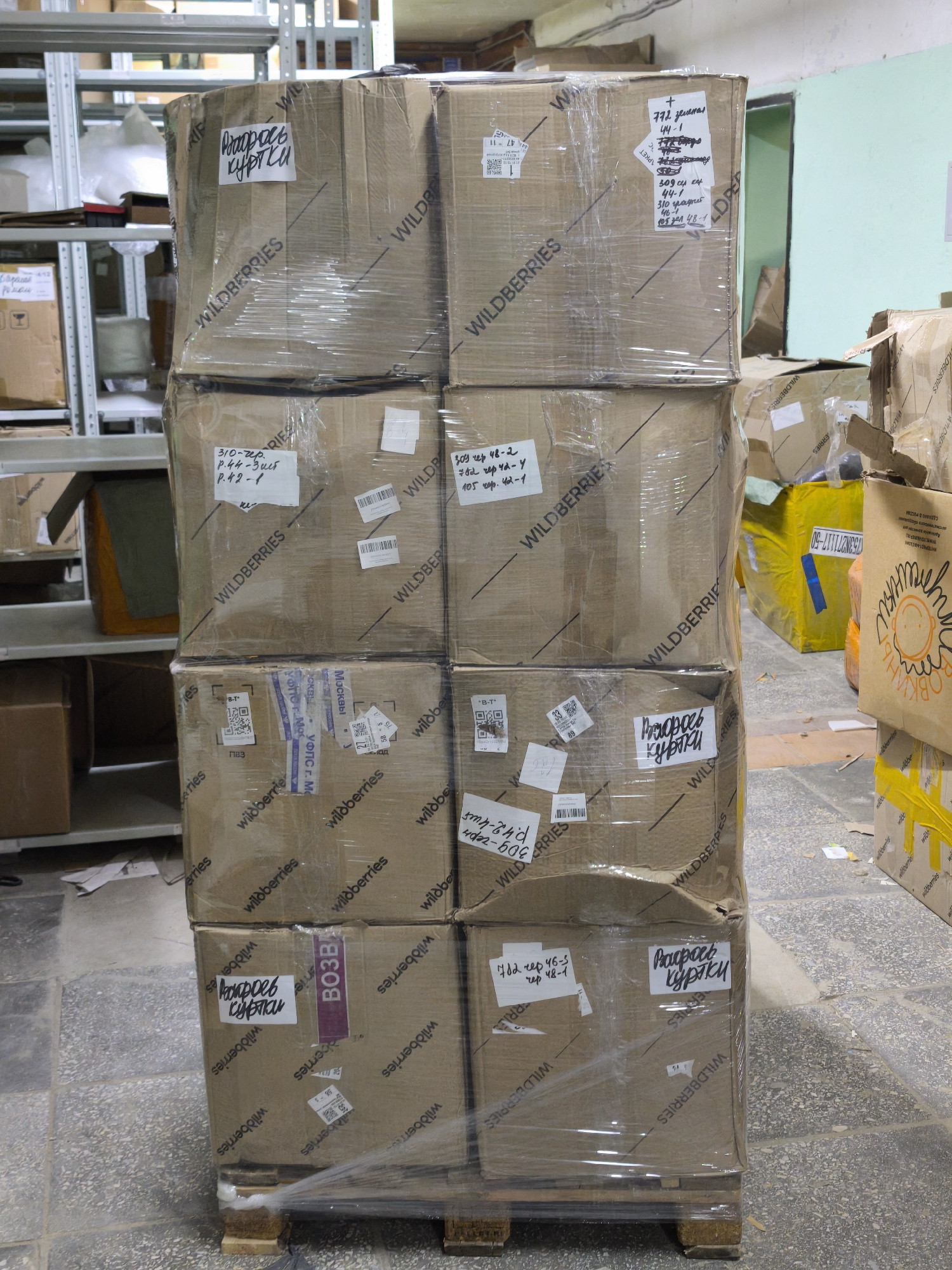 A stack of nine cardboard boxes with the "Wildberries" logo is shrink-wrapped onto a wooden pallet. Each box bears labels, some handwritten in Russian, and other shipping labels, including one mentioning "Moscow". Text on the labels includes dimensions. The boxes are in a warehouse setting with metal shelving visible in the background. Other packages are present nearby, including one with a yellow sticker. The boxes contain "jackets".