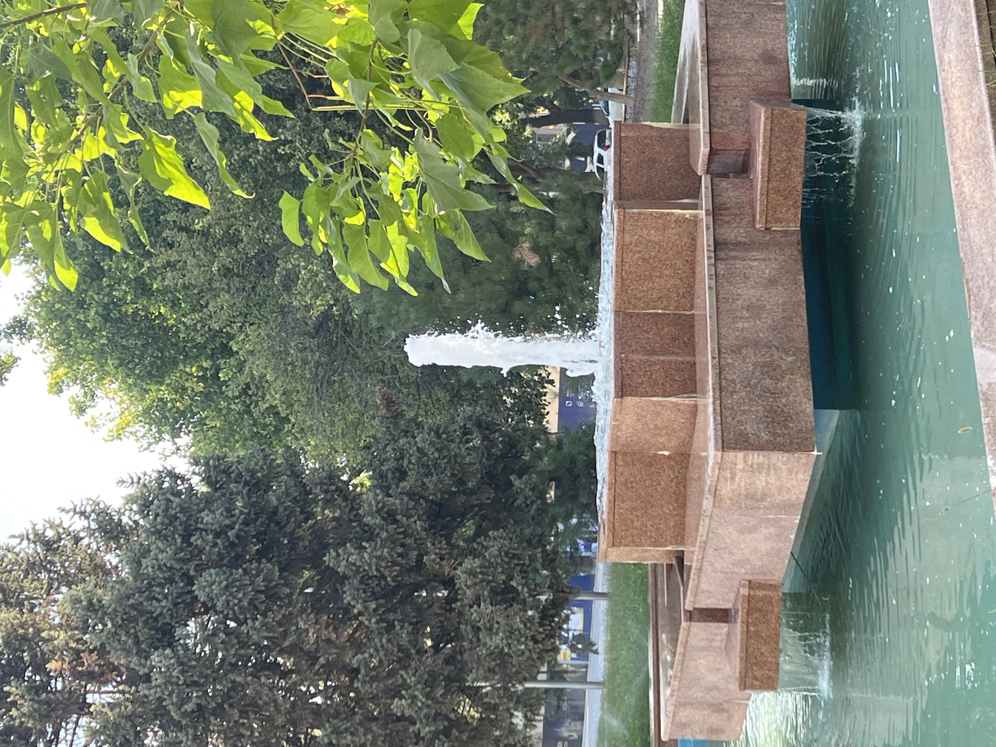A tiered fountain with a central water jet is the main subject of this outdoor scene. The fountain's structure is made of a reddish-brown stone. Water flows from the upper tiers into the lower basin, which is filled with greenish-blue water. Lush green trees and foliage surround the fountain, with some of the trees appearing to be coniferous. In the background, a few white cars are visible parked on a street. The sky is bright, suggesting it is daytime.