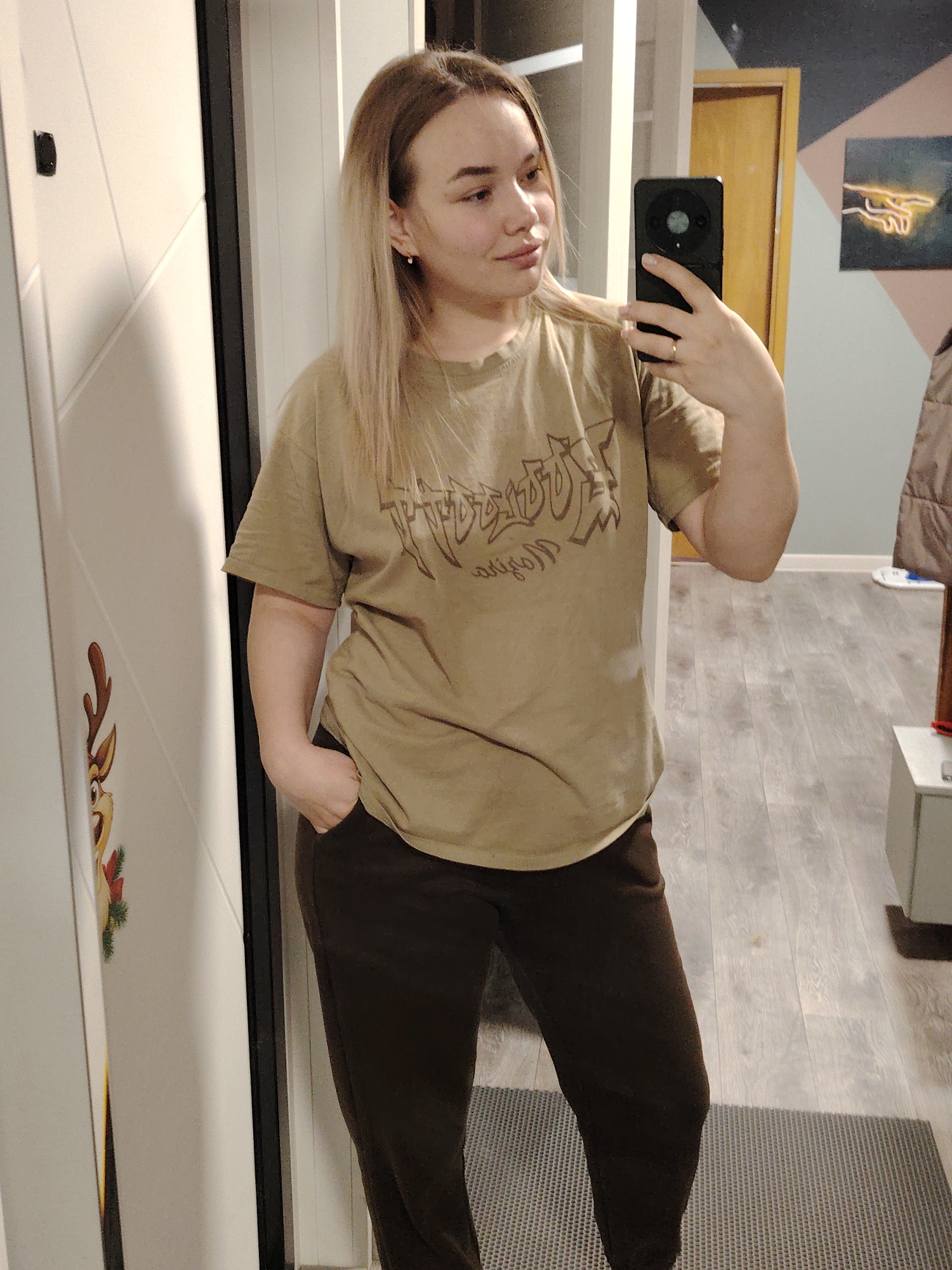 A young woman with blond hair takes a selfie in a hallway. She is wearing a khaki-colored t-shirt with a logo and dark brown sweatpants. Her left hand is in her pocket, and her right hand holds a smartphone. The hallway has light gray wood-look flooring. To the left is a white door with a reindeer decoration. A partially visible open doorway leads to another room with artwork on the wall. The location is Марковский, Russia.