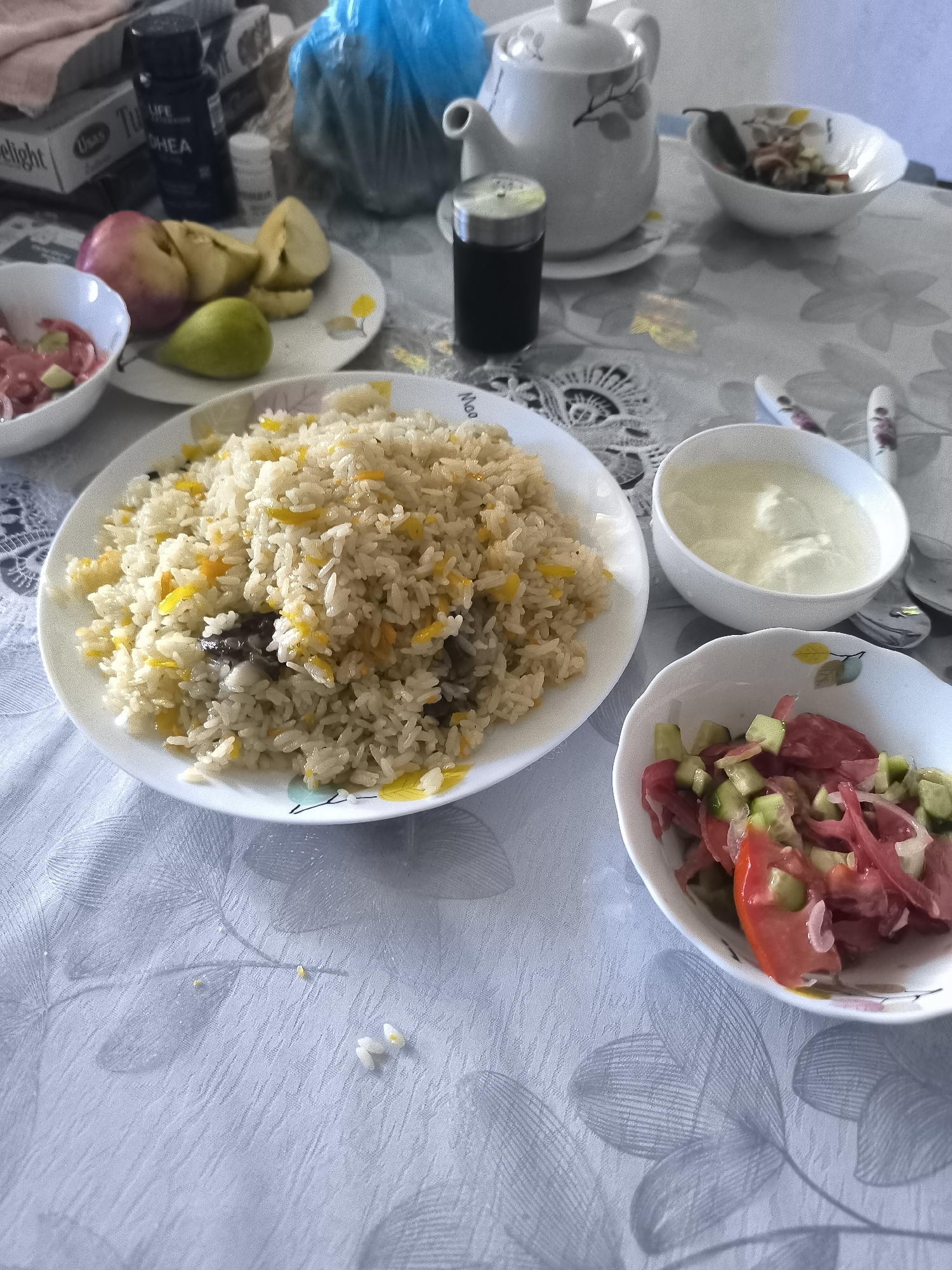 The image shows a table set with food, likely a meal. In the foreground, there is a plate of rice pilaf with what appears to be meat mixed in, and some yellow and orange flecks, possibly spices or vegetables. To the right of the pilaf, there are two small white bowls. One contains a white liquid, possibly yogurt or a sauce. The other bowl holds a salad made of chopped tomatoes, cucumbers, and red onions. Behind the rice plate, there is another plate with sliced apples and a green pear. Further in the background, a white teapot is visible, along with a salt and pepper shaker and other items on the table which are out of focus. The table is covered with a light-colored tablecloth with a floral pattern. The overall impression is a home-style meal setting in Kokand, Uzbekistan, characterized by fresh ingredients and traditional dishes.