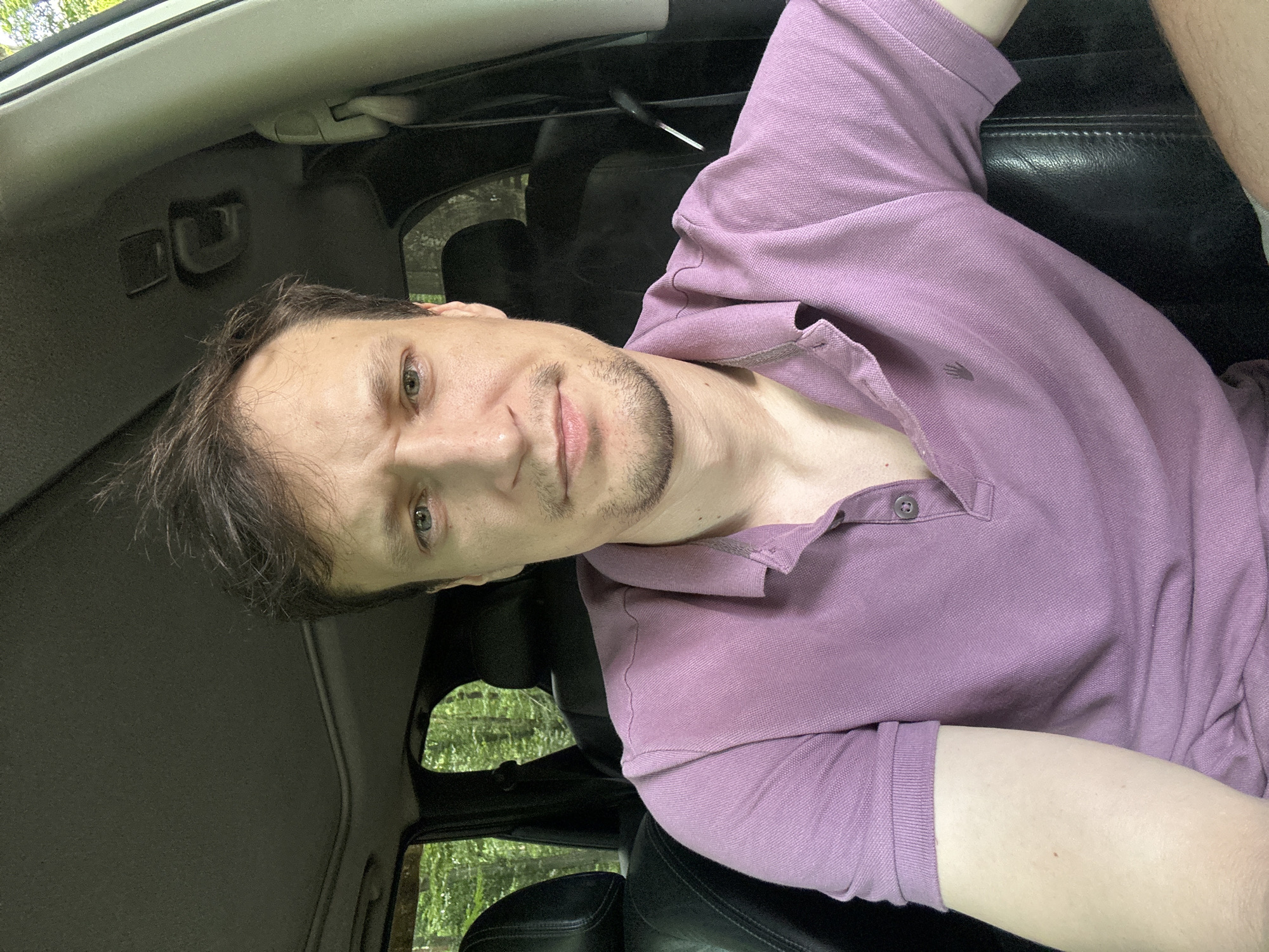A medium shot photo shows a light-skinned man with brown hair sitting in the driver's seat of a car. He is looking directly at the camera with a neutral expression. He is wearing a short-sleeved, purple polo shirt with two buttons at the collar. The interior of the car is visible, including the black leather seats and the sunroof. Outside the car, green trees are visible through the windows, suggesting a natural or wooded environment, possibly on the outskirts of Shatava, Ukraine. The image is well-lit, likely taken during the daytime.