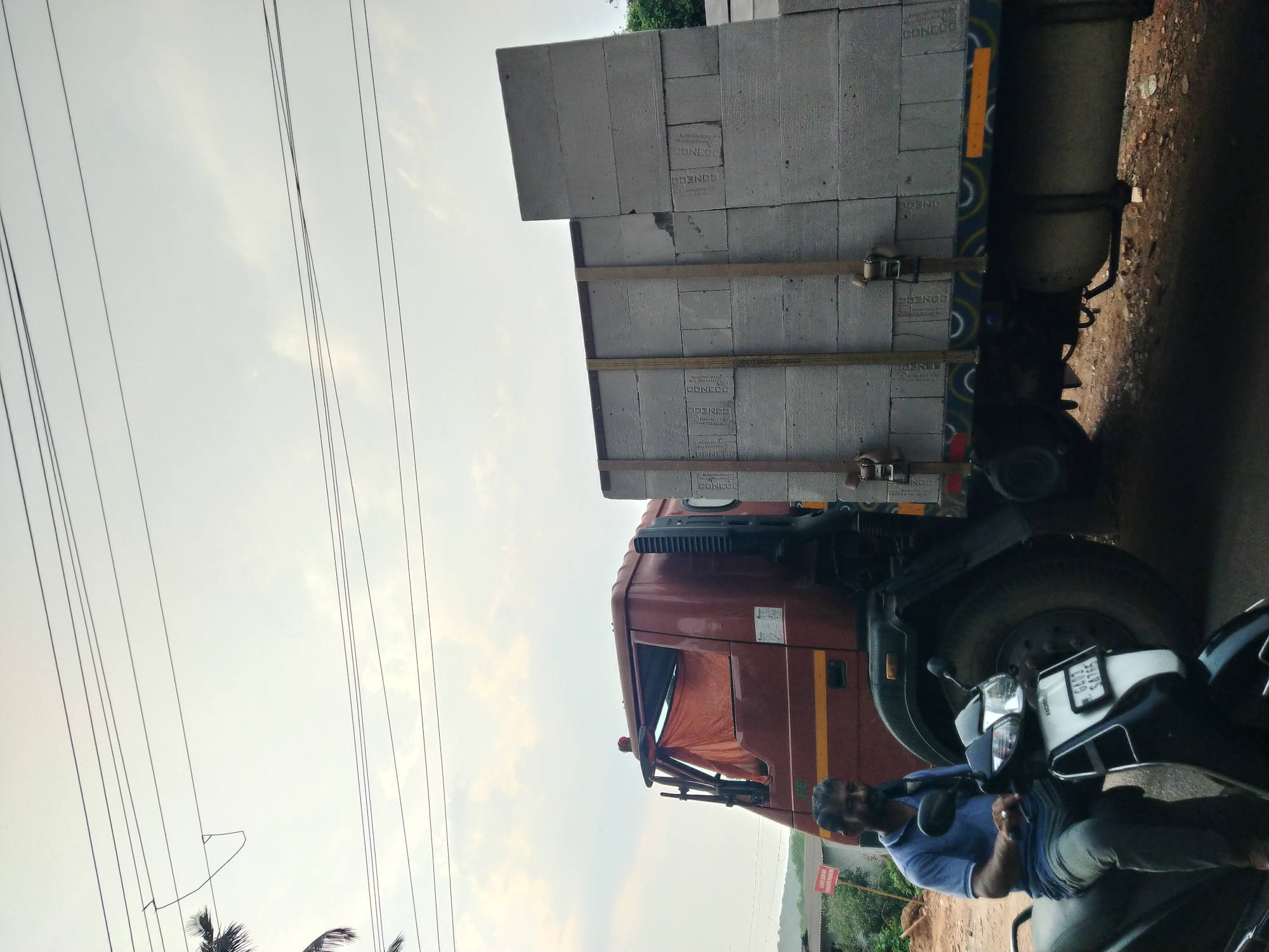 The image captures a scene alongside a road in Oxel, India. In the foreground, a man is seated on a parked scooter. Beside the scooter is a truck loaded with concrete blocks, stacked high. The blocks are branded with the word "CONECCO". The truck is parked on the side of the road, on an unpaved shoulder. Above, there is a sky that's getting to be evening. Power lines run horizontally across the upper left portion of the frame, creating a visual contrast with the natural elements. In the background, there is green vegetation. The scene appears to be an everyday occurrence.