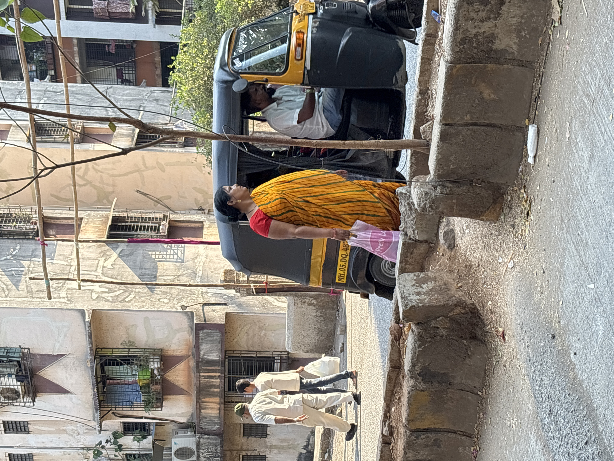The image captures a street scene in Ambernath, India. In the foreground, a woman wearing a yellow and orange sari with a red blouse is seen walking on the street, carrying a pink plastic bag. Adjacent to her, a black and yellow auto-rickshaw is parked, with its driver inside. Behind the woman and auto-rickshaw, two men wearing white uniforms are walking. The background features apartment buildings.