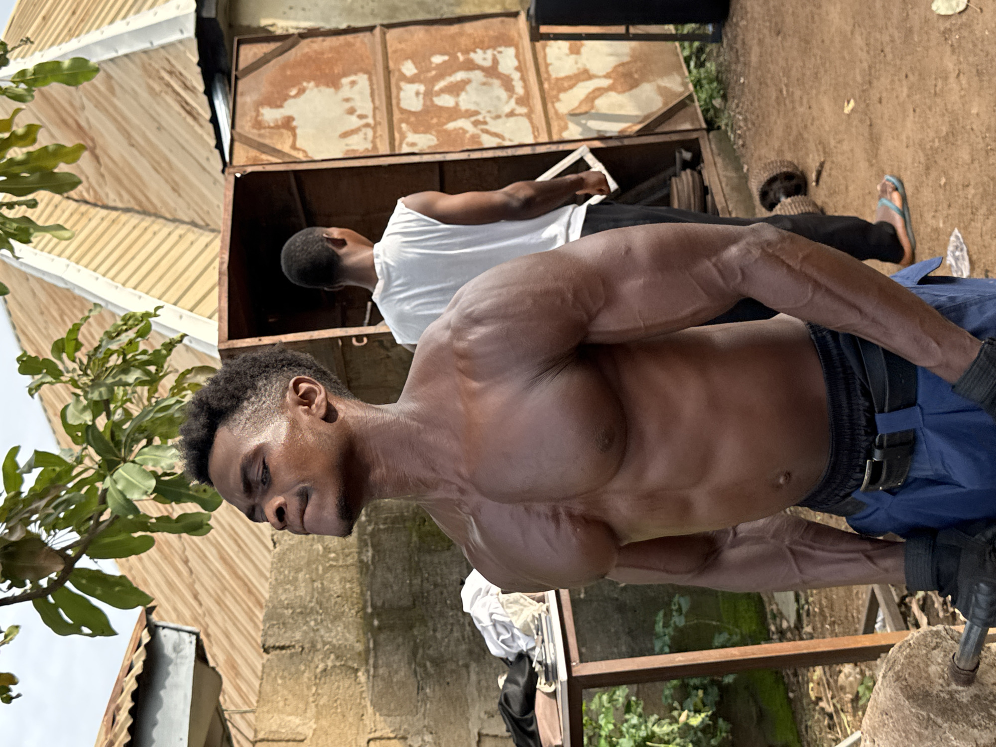 In Akwanga, Nigeria, a muscular Black man with dark skin and short, dark, curly hair is shown from the chest up, looking to his left with his head tilted back and eyes closed. He has prominent pectoral muscles, deltoids, and biceps, with visible veins on his arms. He is wearing black gloves, a black belt with a silver buckle, and blue trousers. To the right of the main subject, another Black man with short, dark hair is partially visible from the back, wearing a white sleeveless t-shirt and black trousers. Behind them, a dilapidated, rust-colored metal structure with an open door is situated in a dusty, outdoor environment. A table with some folded clothes is visible to the left. The background shows a rough stone wall and a corrugated metal roof. The ground is made of dirt and scattered leaves.