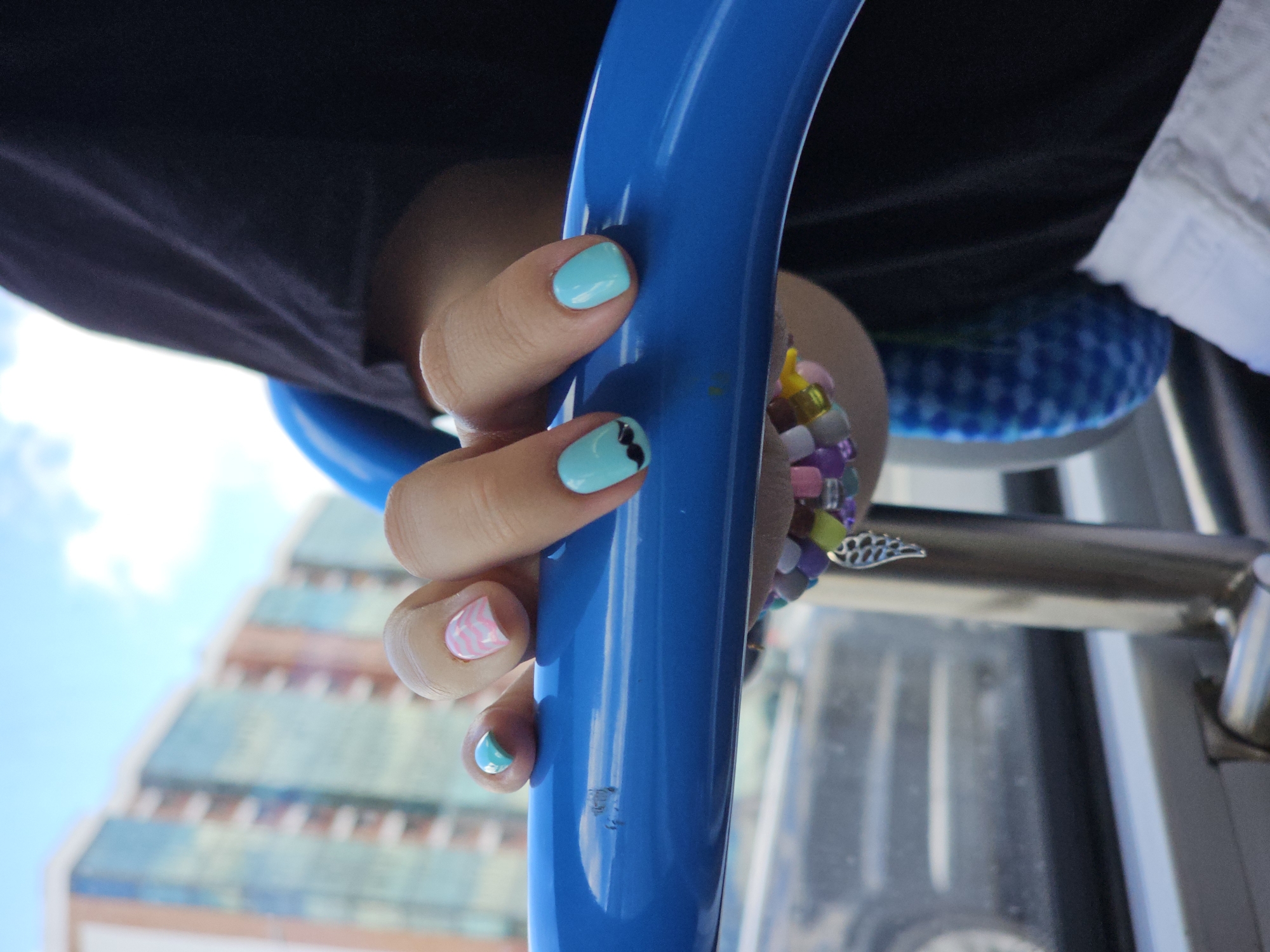 In Novoivanovskoe, Russia, a close-up shot focuses on a person's hand holding onto a bright blue bar. The individual has painted fingernails in various colors and designs: one nail has a pink zigzag pattern, another is a solid teal, a third is teal with a black mustache design, and the thumb has a light pink and white zigzag pattern. Attached to the hand is a beaded bracelet with colorful beads, a silver wing charm, and a blue and white checkered fabric accessory. In the background, a modern building with glass windows is visible, reflecting the sky. The scene appears to be taken from inside a vehicle, as suggested by the presence of a car window and part of a car's interior. The sky is clear with scattered clouds.