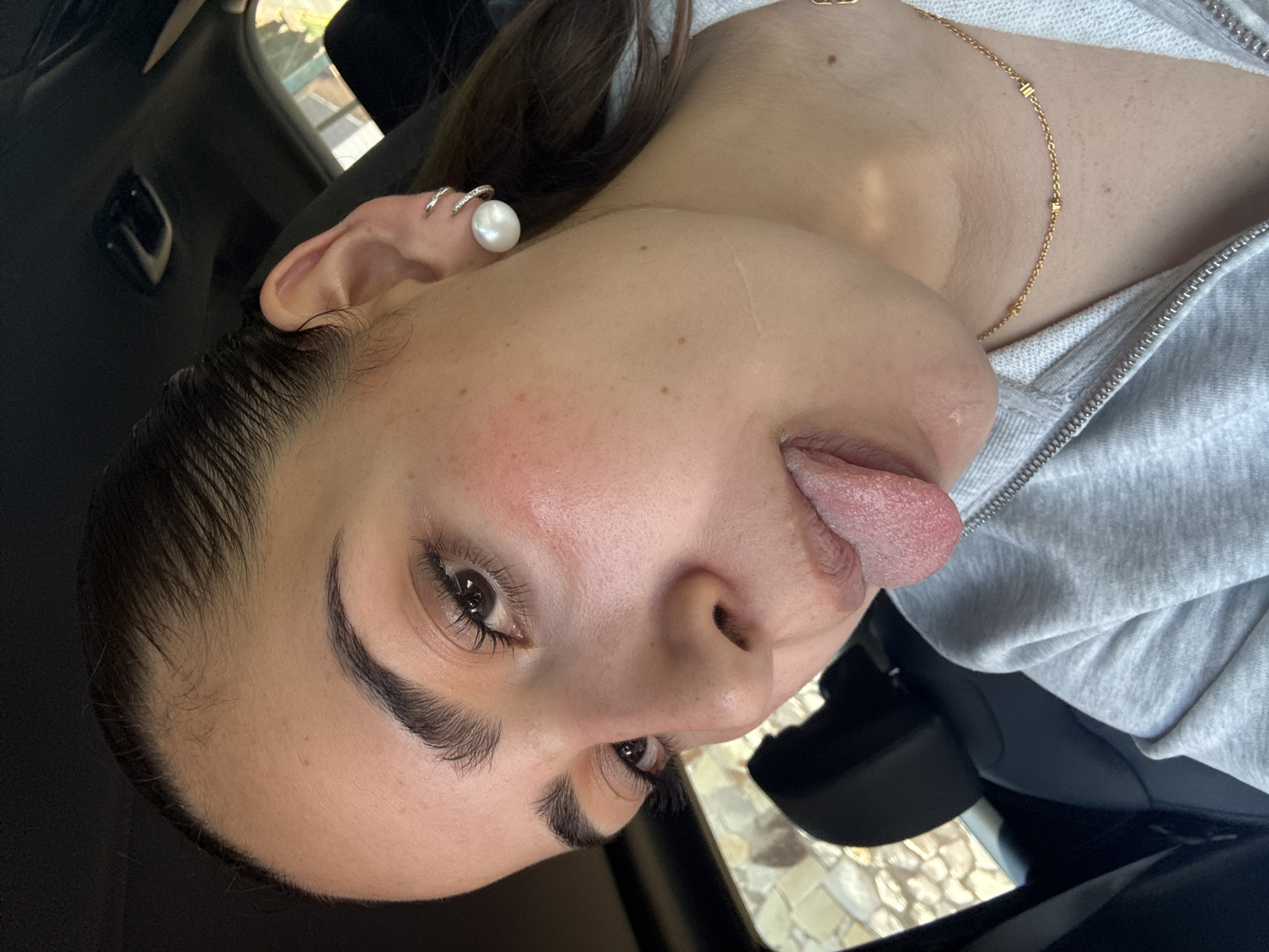A close-up, tilted portrait shows a young woman playfully sticking out her tongue, captured likely inside a vehicle during the daytime in San Bernardino Tlaxcalancingo, Mexico. Her head is angled to the left, and she looks directly at the camera with a casual, expressive demeanor.
The woman has dark hair, which appears to be wet or slicked back at the hairline. Her eyebrows are dark and well-defined, and she has visible eyelashes. Her skin shows some natural freckles and a slight flush on her cheek. In her left ear, she wears two earrings: a prominent, large white pearl stud and a smaller, possibly silver-toned ring or cuff above it.
She is wearing a light grey hooded sweatshirt with a visible zipper down the front. Around her neck, a delicate gold chain necklace with small, rectangular or square-shaped beads is visible. The background suggests the interior of a car, with dark upholstery or plastic surfaces visible behind her head and shoulders. Through what appears to be a window, some outdoor light and a hint of light-colored, possibly paved ground or a building exterior are visible, indicating daylight. No text is visible in the image.