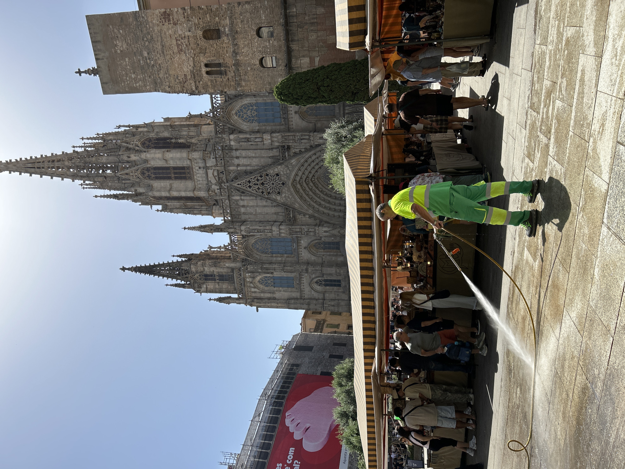 In Barcelona, Spain, in front of the Barcelona Cathedral, a street cleaner in a neon green and yellow uniform is hosing down the stone pavement. The cleaning is taking place amidst an outdoor market with numerous stalls covered by striped awnings, and a crowd of people browsing the merchandise. The Barcelona Cathedral, a grand Gothic structure with intricate spires and stained-glass windows, dominates the background. To the left, a large building is under construction, draped with a red banner featuring an advertisement. The sky is clear and blue, indicating a sunny day.