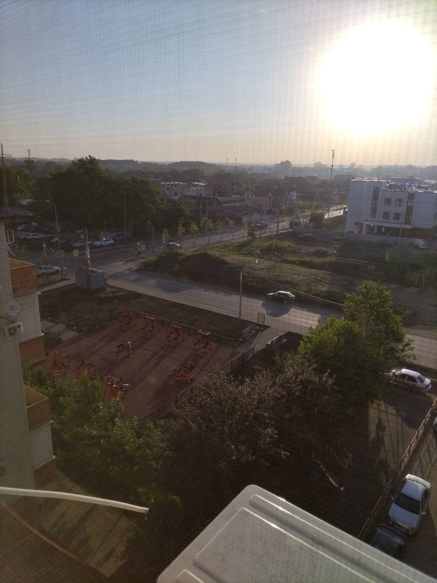 An aerial shot captures a suburban street scene in Rossiyskiy, Russia, bathed in the warm, low light of a setting or rising sun. The sky is a pale blue, transitioning to a hazy yellow near the bright, almost blinding sun positioned in the upper right quadrant. In the foreground, the top of a light-colored building with a textured surface is visible, partially obscuring the view.
Below, a red-surfaced outdoor exercise area is laid out with various pieces of red gym equipment, casting long shadows. A single individual is visible on the exercise equipment, appearing to be engaged in a workout. Surrounding this area is dark fencing and lush green trees.
Further into the scene, a paved road runs horizontally, with several cars parked along its left side and others driving on it. One dark sedan is in motion on the road. Across the road, a modern white building with multiple windows stands prominently. Beyond these elements, a sprawling landscape of smaller houses with red roofs and dense green trees stretches towards the horizon, with some distant structures and a tall tower or antenna visible. The overall impression is one of a peaceful, everyday urban environment under a vast, bright sky.