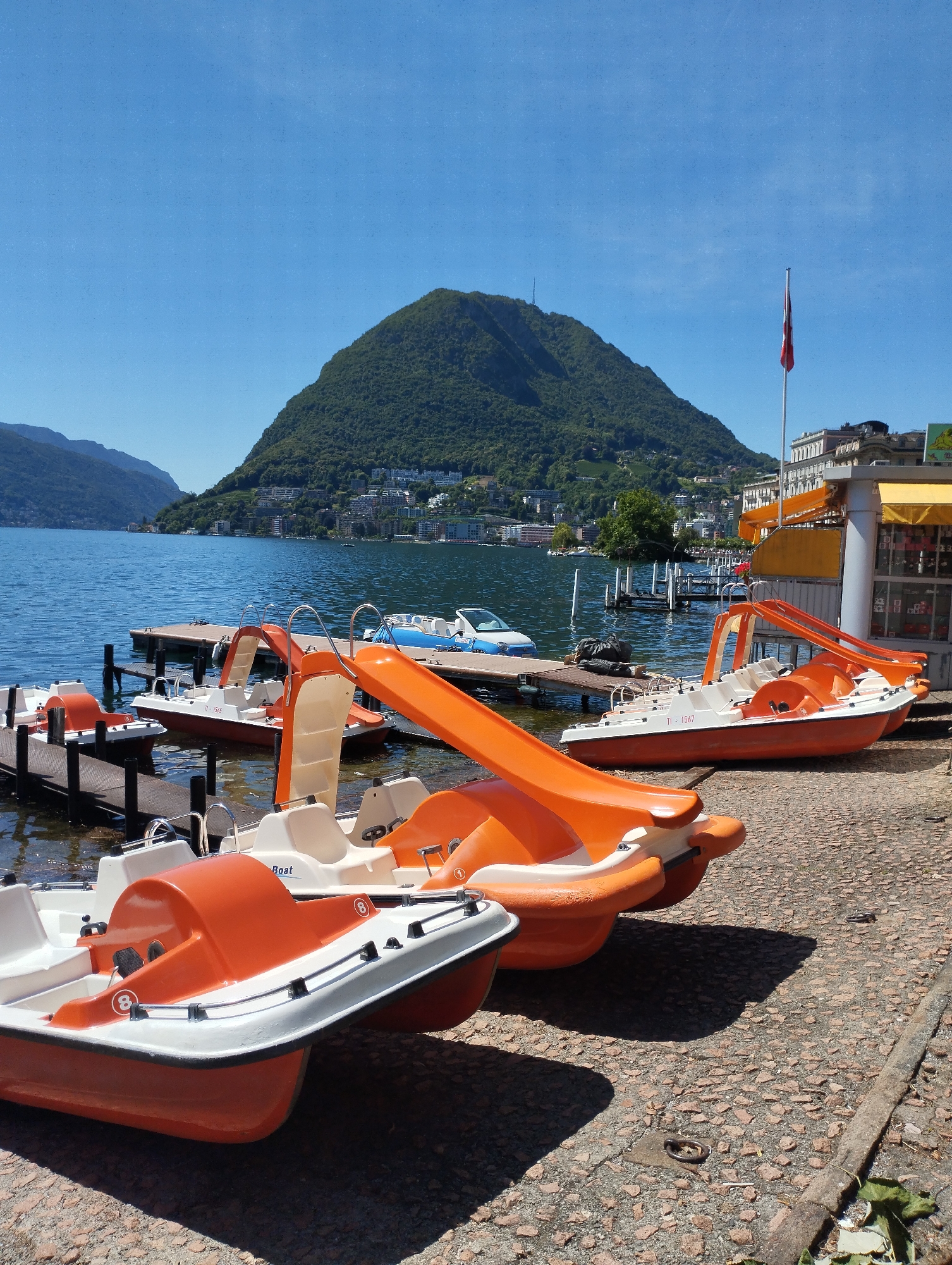 The image depicts a sunny day at the lakefront in Lugano, Switzerland. Several orange and white pedal boats are lined up on a gravel shore, some partially in the water, attached to wooden docks. A prominent green, tree-covered mountain rises in the background, with buildings visible on its slopes and at its base. A Swiss flag flies from a flagpole atop a building on the right side of the frame. The lake is calm and blue, reflecting the clear sky. There are no people visible in the image.